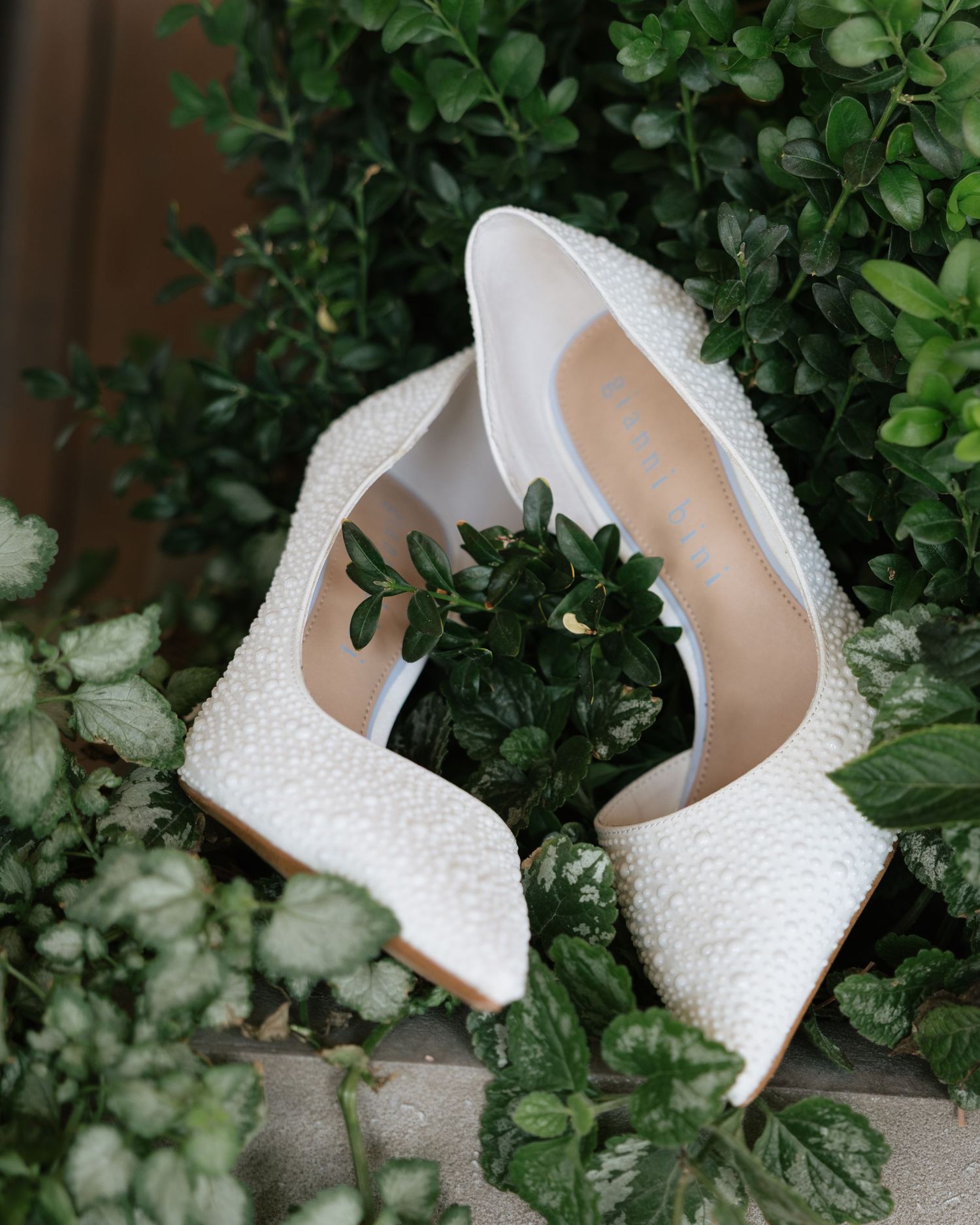 Pearl-embellished white bridal heels displayed among lush green garden foliage.