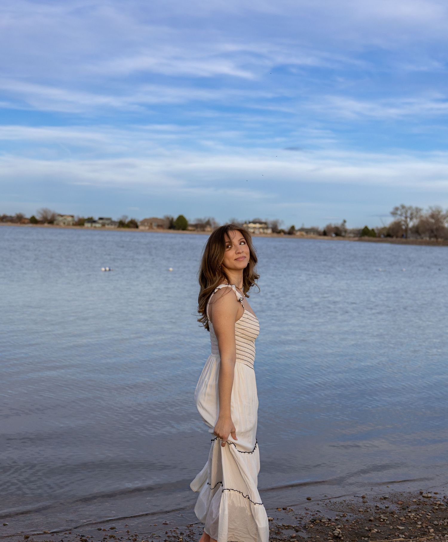 Young woman in white dress standing by a lake shore under a blue cloudy sky.