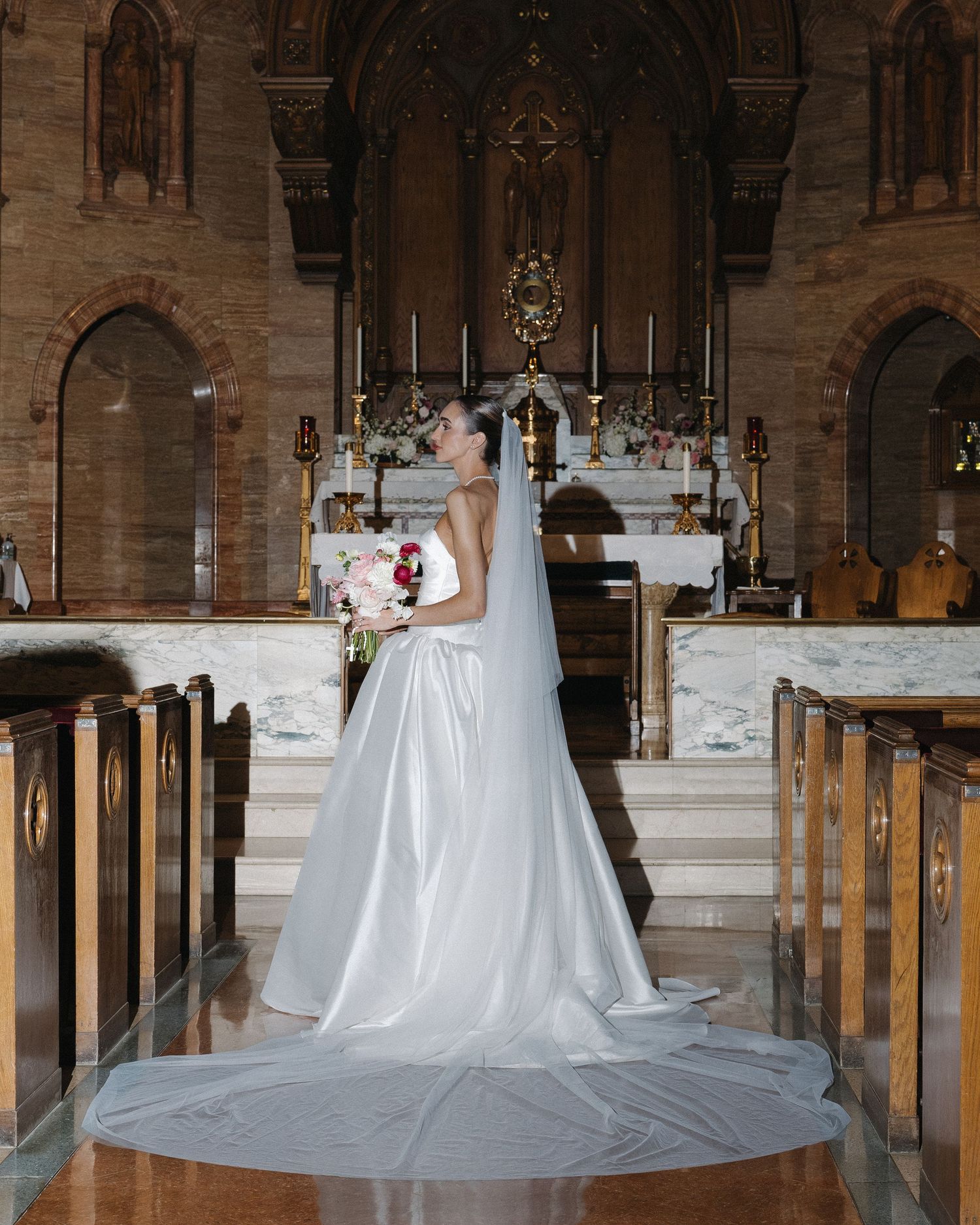 Bride in a white gown with cathedral veil stands in a historic church aisle holding a bouquet.