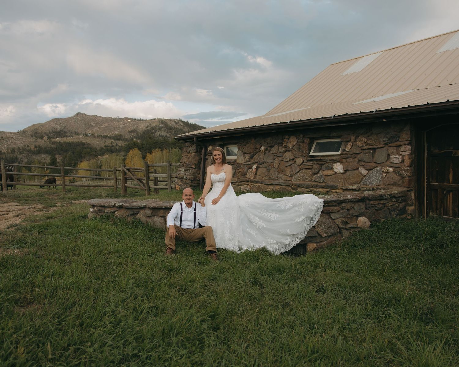 Bride and groom pose outside rustic stone barn with mountain backdrop on their wedding day.