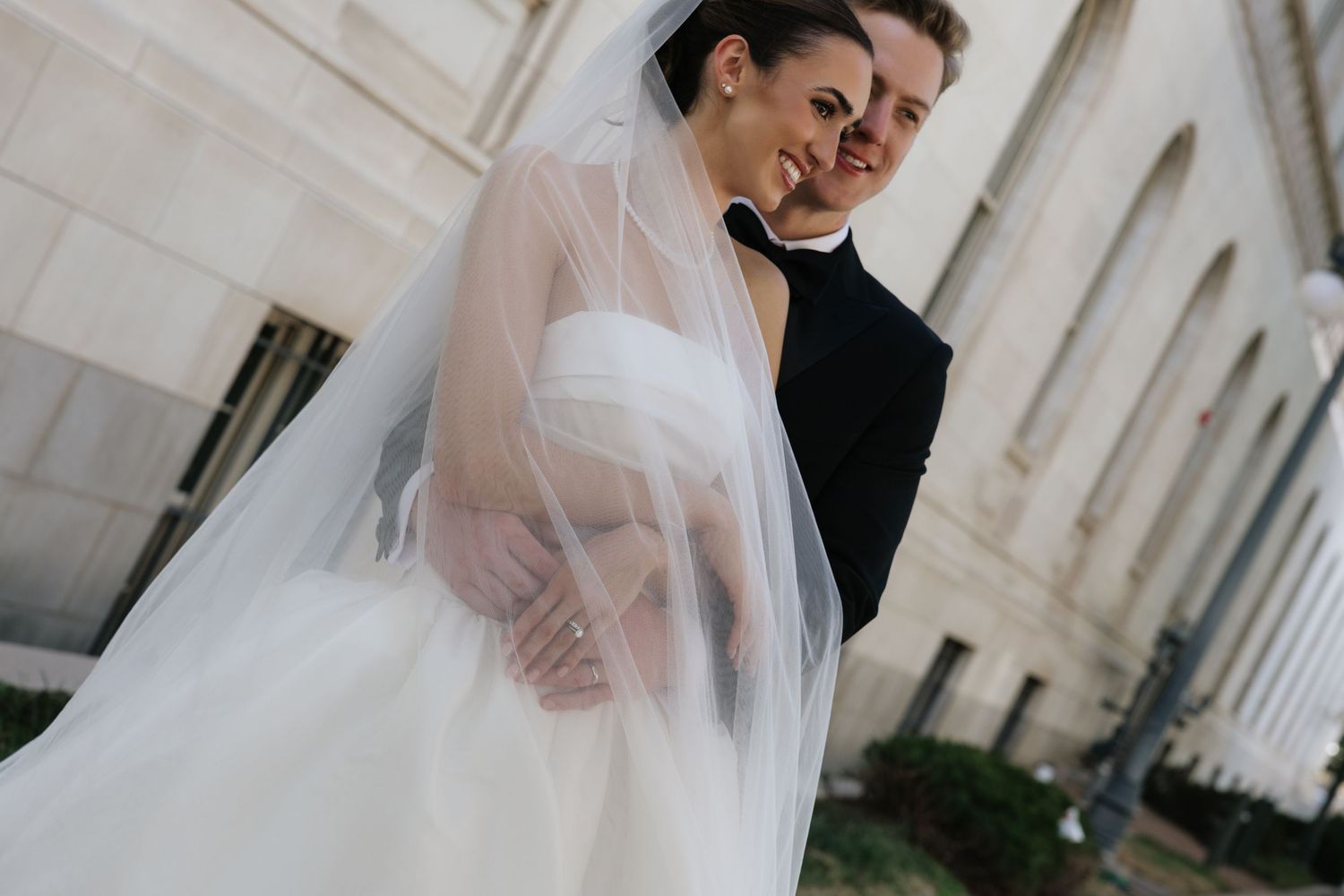 Bride in white ball gown and long veil smiles with groom in black suit outside stone building.