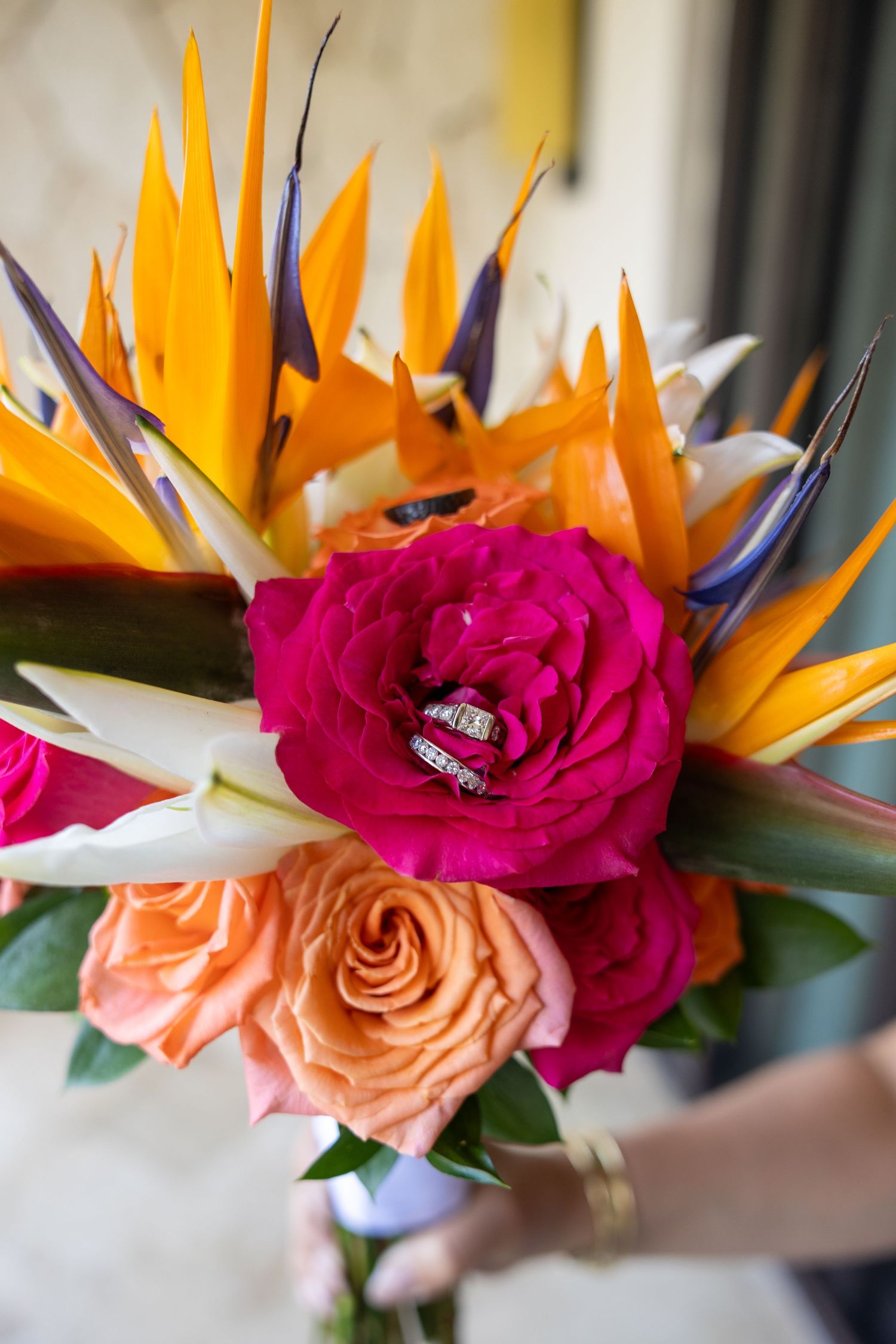 Tropical bridal bouquet with bird of paradise, hot pink and orange roses with wedding rings.