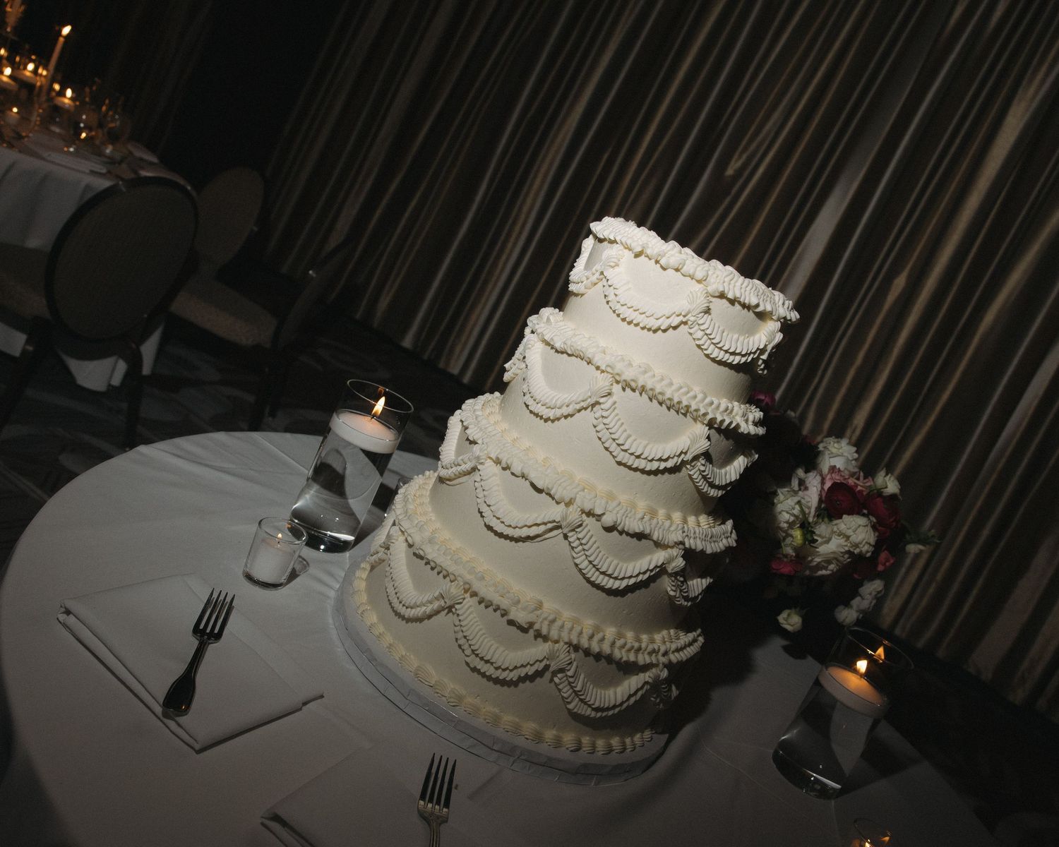 Elegant four-tier white wedding cake with ruffle and pearl decorations displayed on reception table.