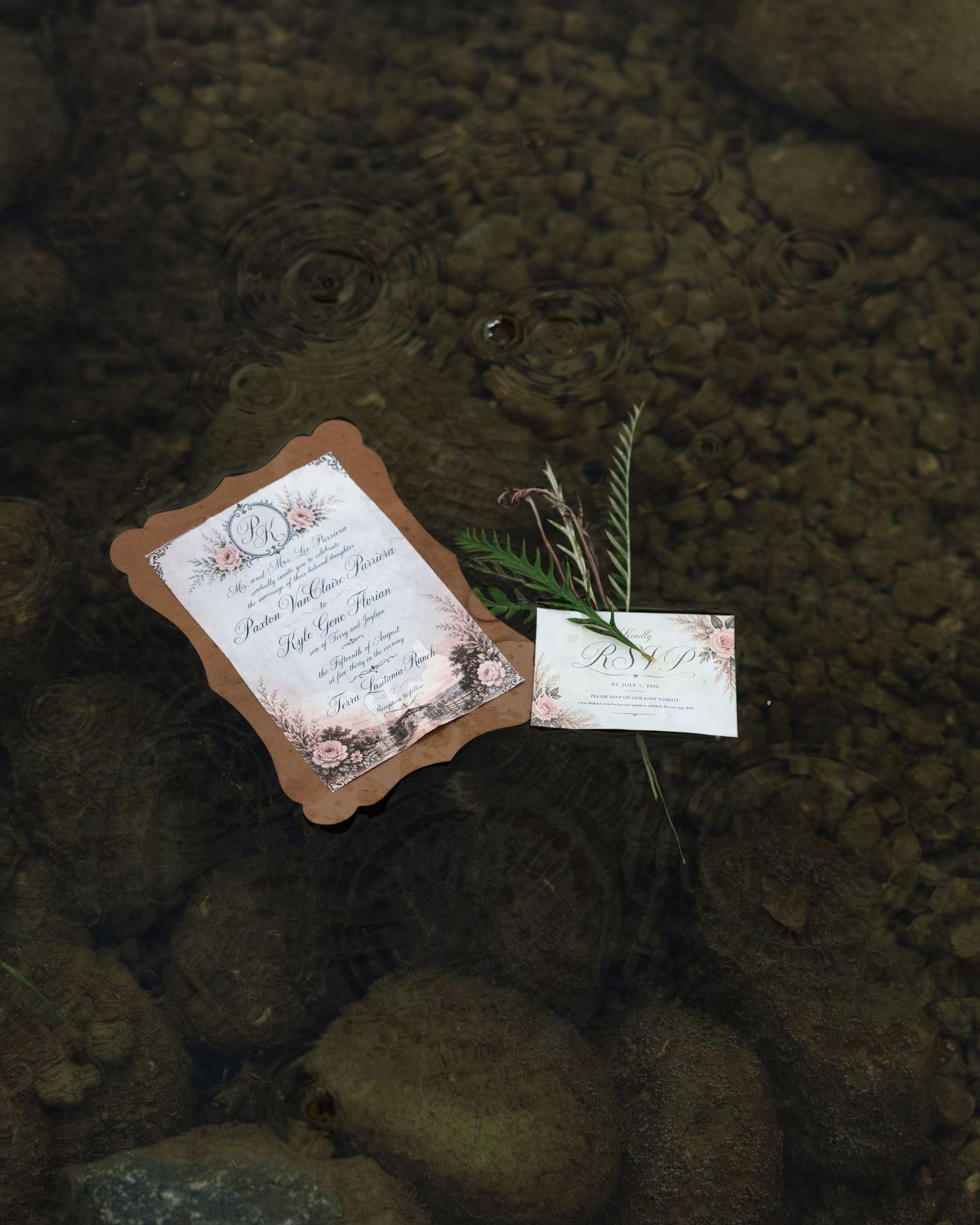 Floral wedding invitation suite with kraft paper border resting on smooth river rocks in shallow water.