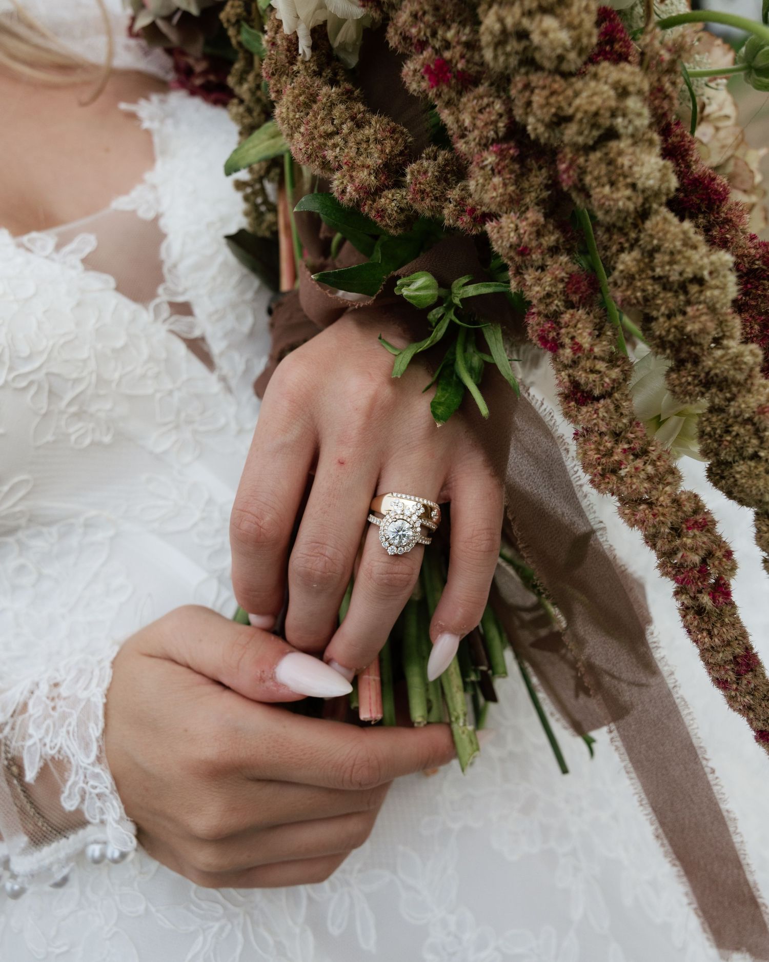 Bride wearing diamond halo engagement ring holding rustic bouquet with greenery and burgundy flowers.