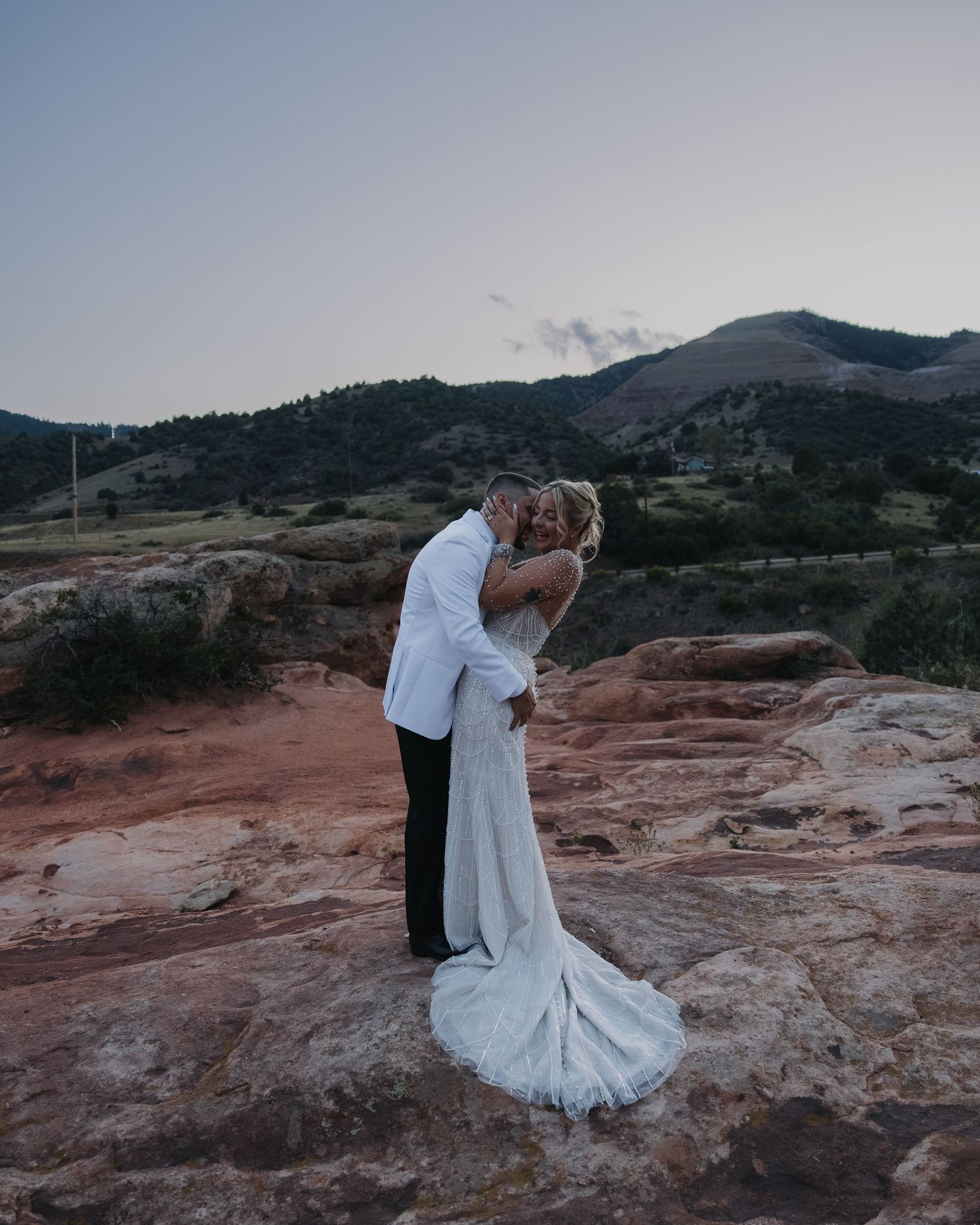 Bride and groom share a kiss on red rock formation with mountain landscape at sunset wedding.
