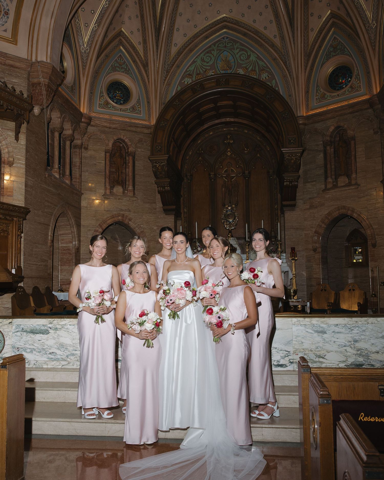 Bride and bridesmaids in blush gowns pose inside ornate Gothic church.