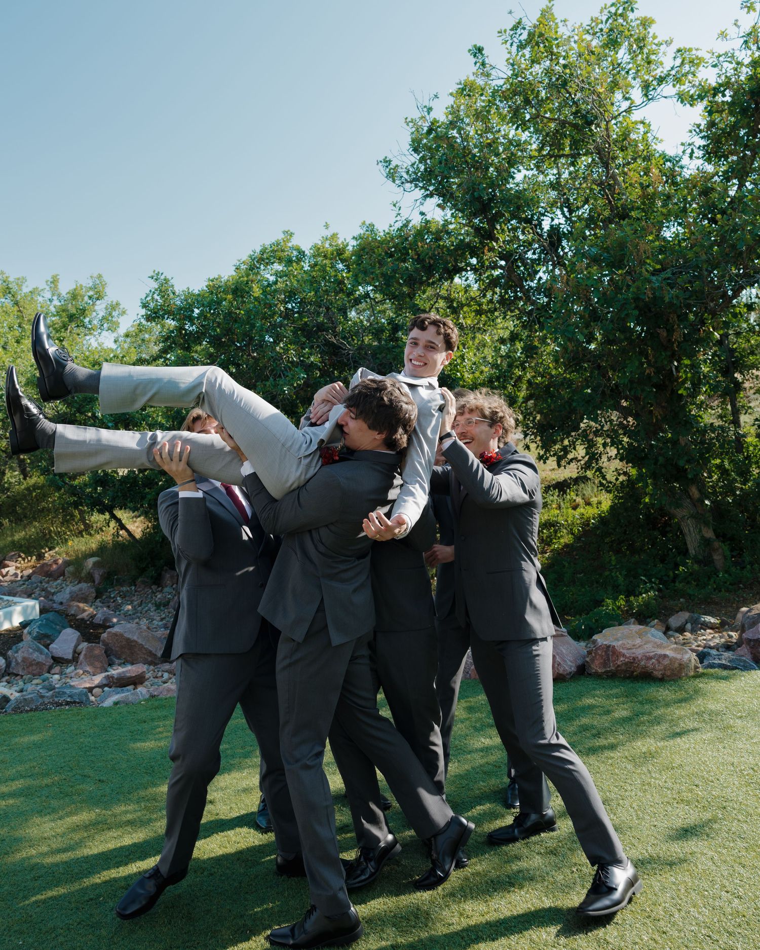 Groomsmen in gray suits playfully lift the groom overhead on a sunny green outdoor lawn.