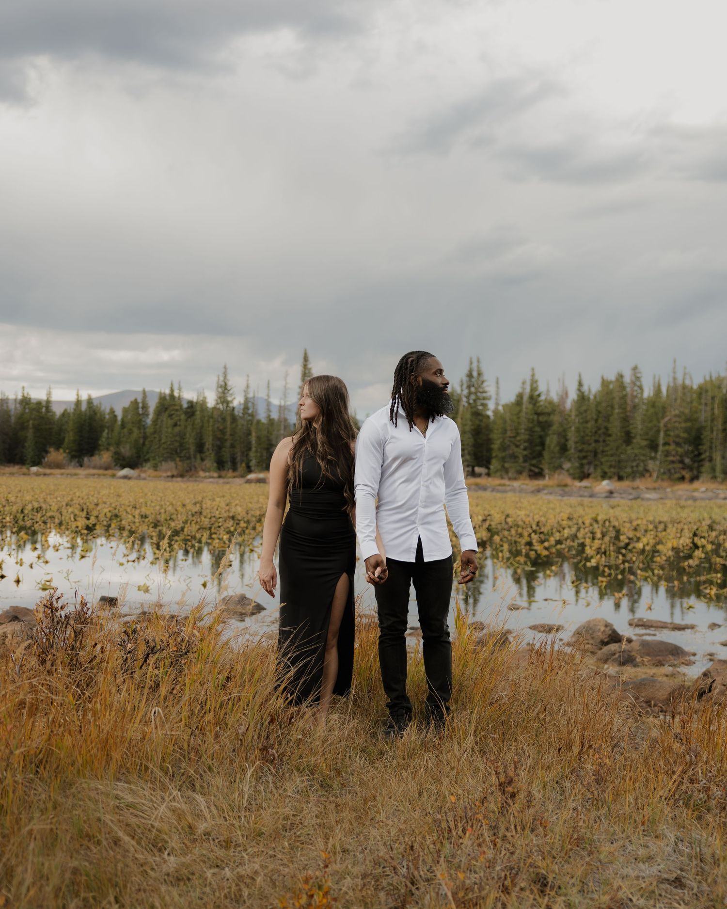 Couple standing hand in hand facing a scenic alpine lake with stormy skies and pine forest.