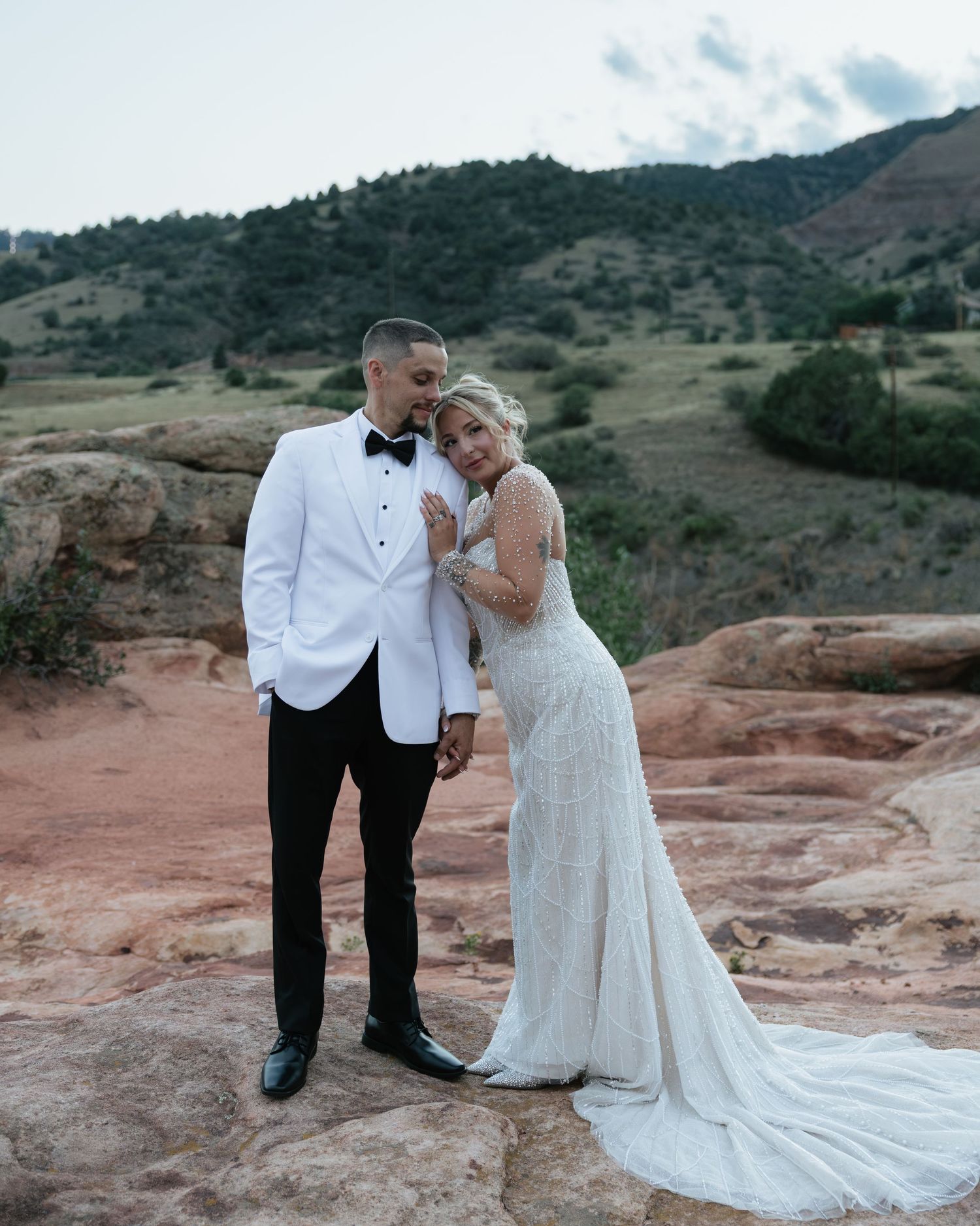 Bride and groom standing on red rock formations, groom in white jacket, bride in beaded train gown.