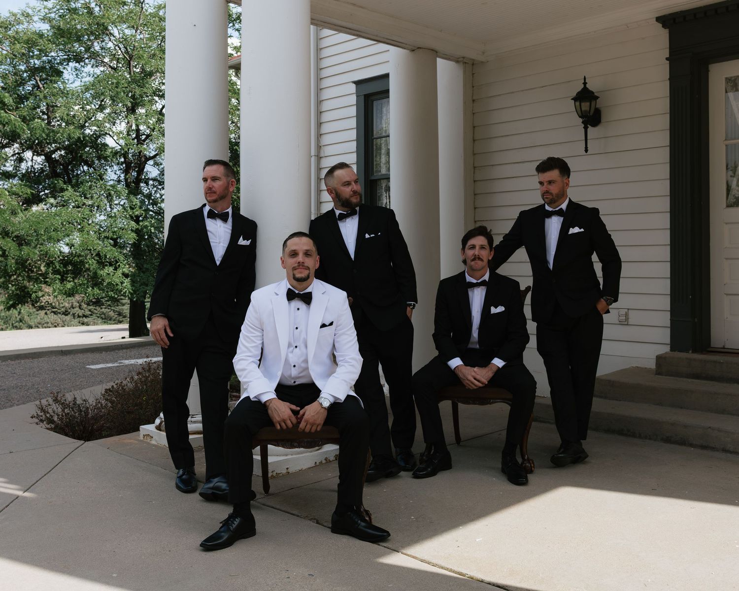 Groom in white tuxedo poses with groomsmen in black tuxedos on white mansion porch.