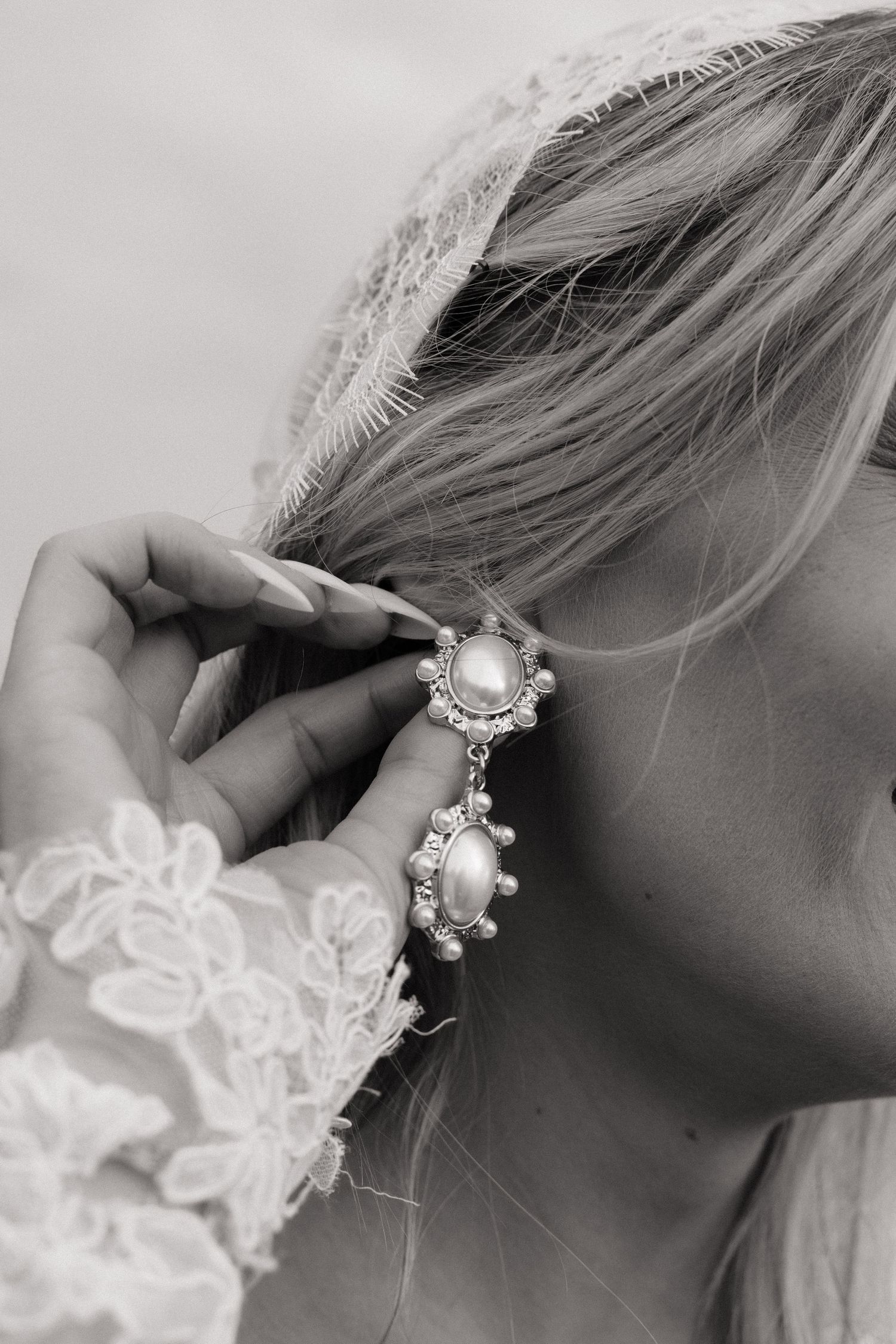Black and white close-up of bride wearing pearl and crystal drop earrings with lace veil and sleeve.