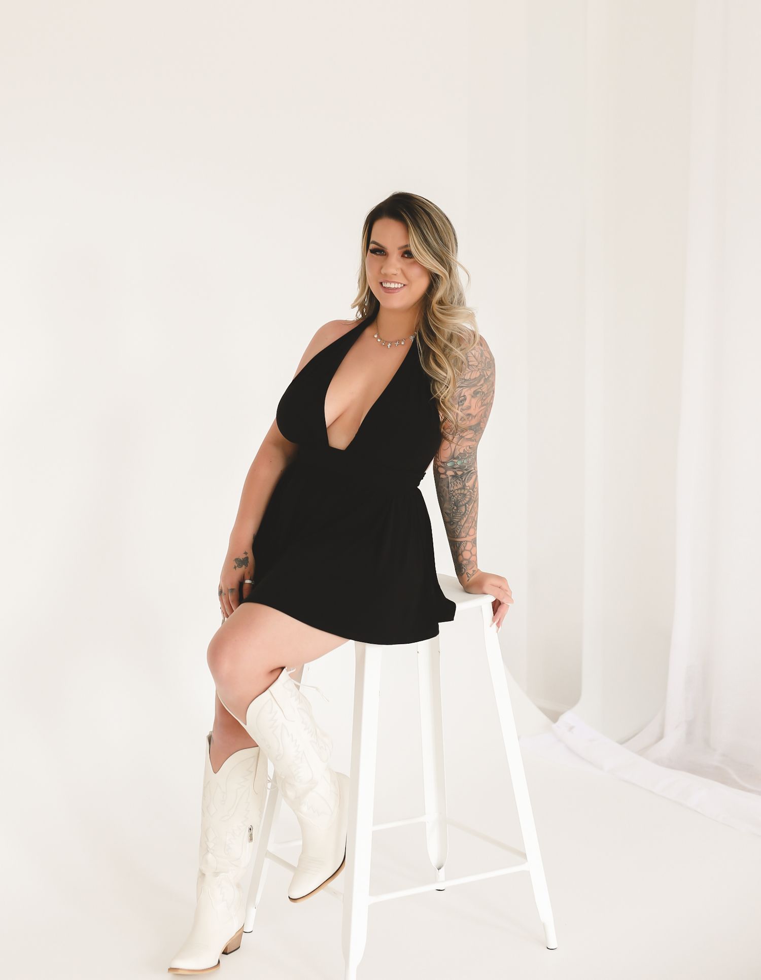 Woman in black halter dress and white cowboy boots poses on a stool against a white backdrop.