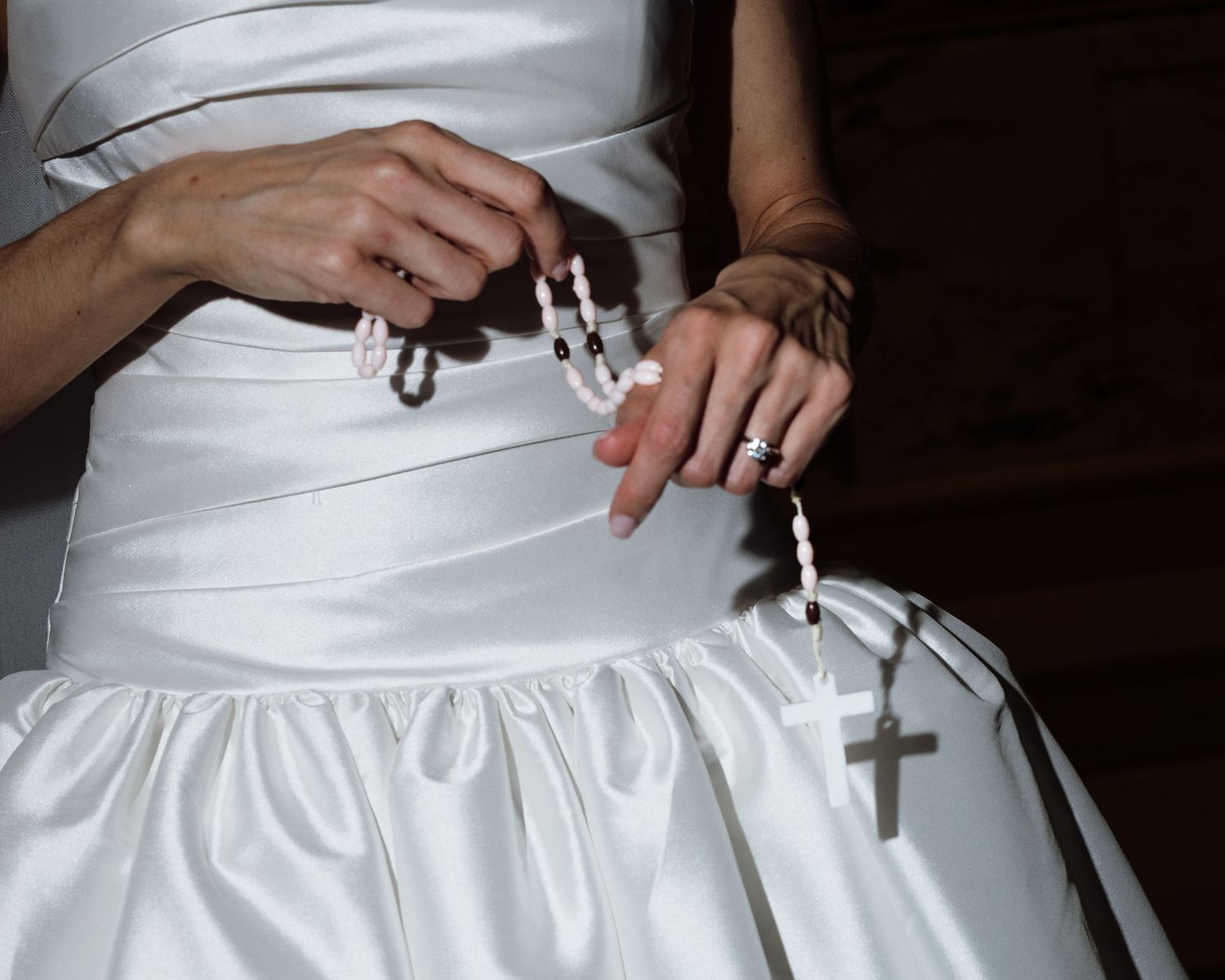 Bride holds rosary beads in white satin wedding gown close-up.