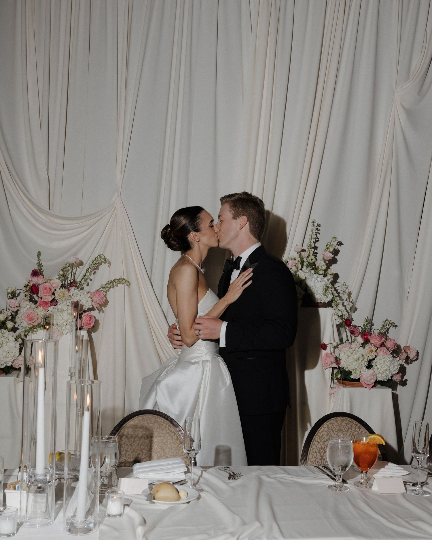 Bride and groom share a kiss at their elegant wedding reception sweetheart table with floral arrangements.
