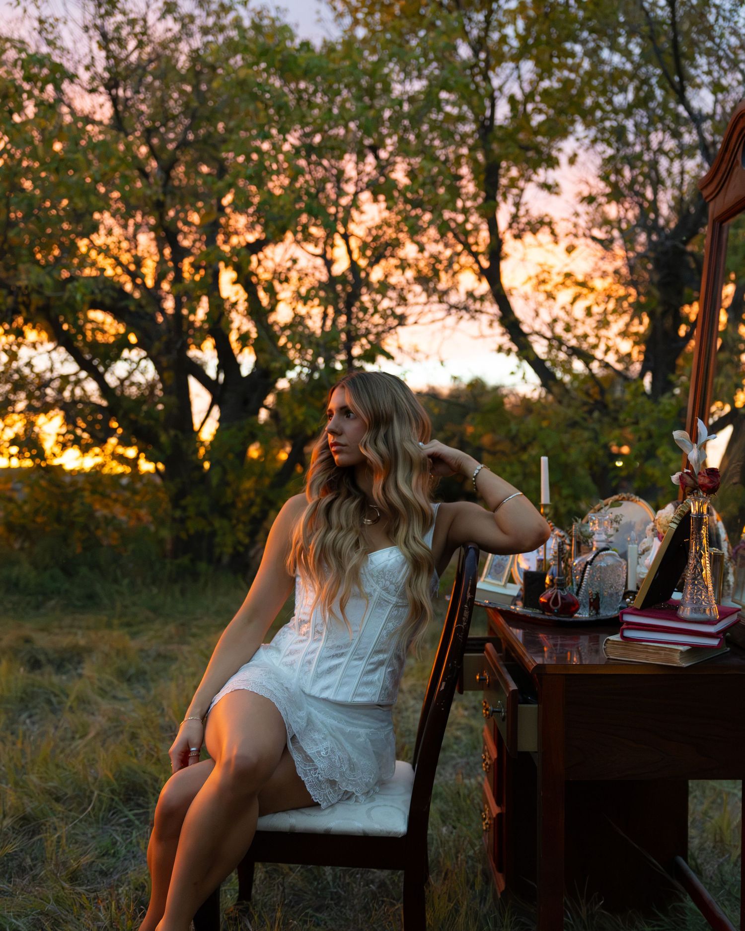 Woman in white lace dress sits at vintage vanity outdoors at sunset with golden trees in background.
