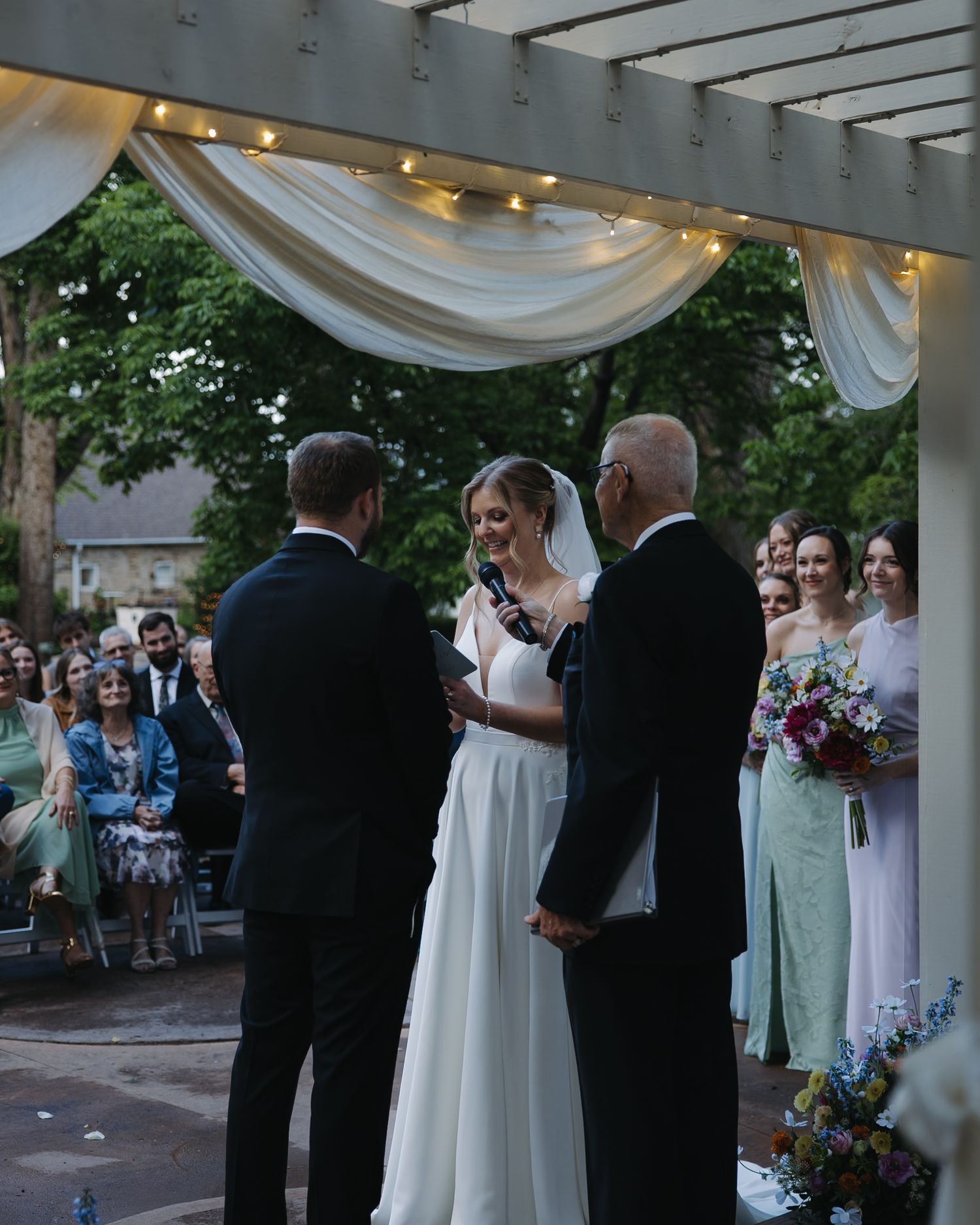 Bride reads vows to groom under draped pergola with string lights at outdoor evening wedding ceremony.