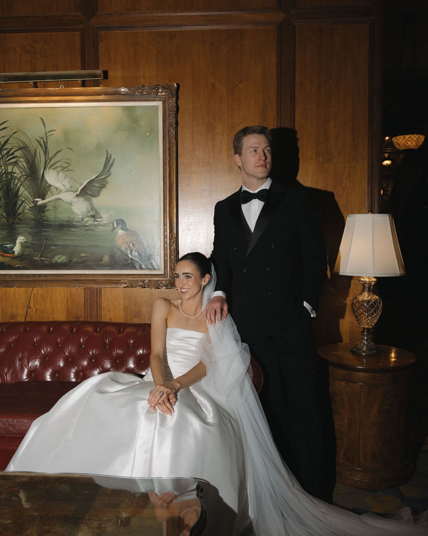 Bride in strapless satin gown and groom in tuxedo pose beside leather sofa in classic wood-paneled room.