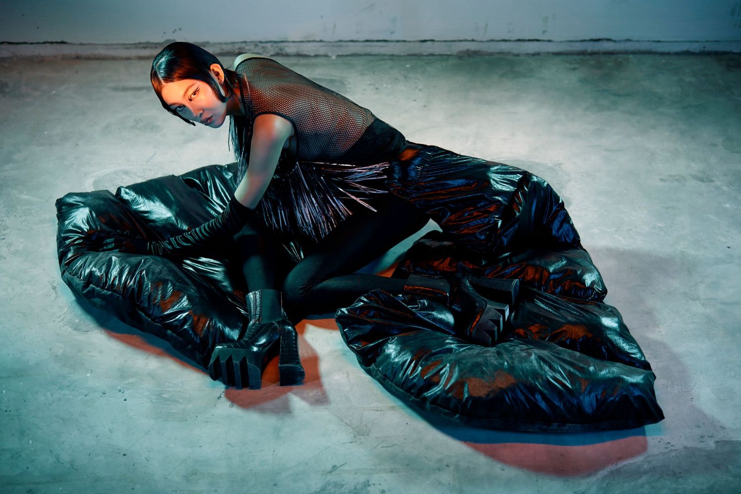 Model reclines on floor wearing glossy black oversized puffer ensemble in editorial fashion shoot.