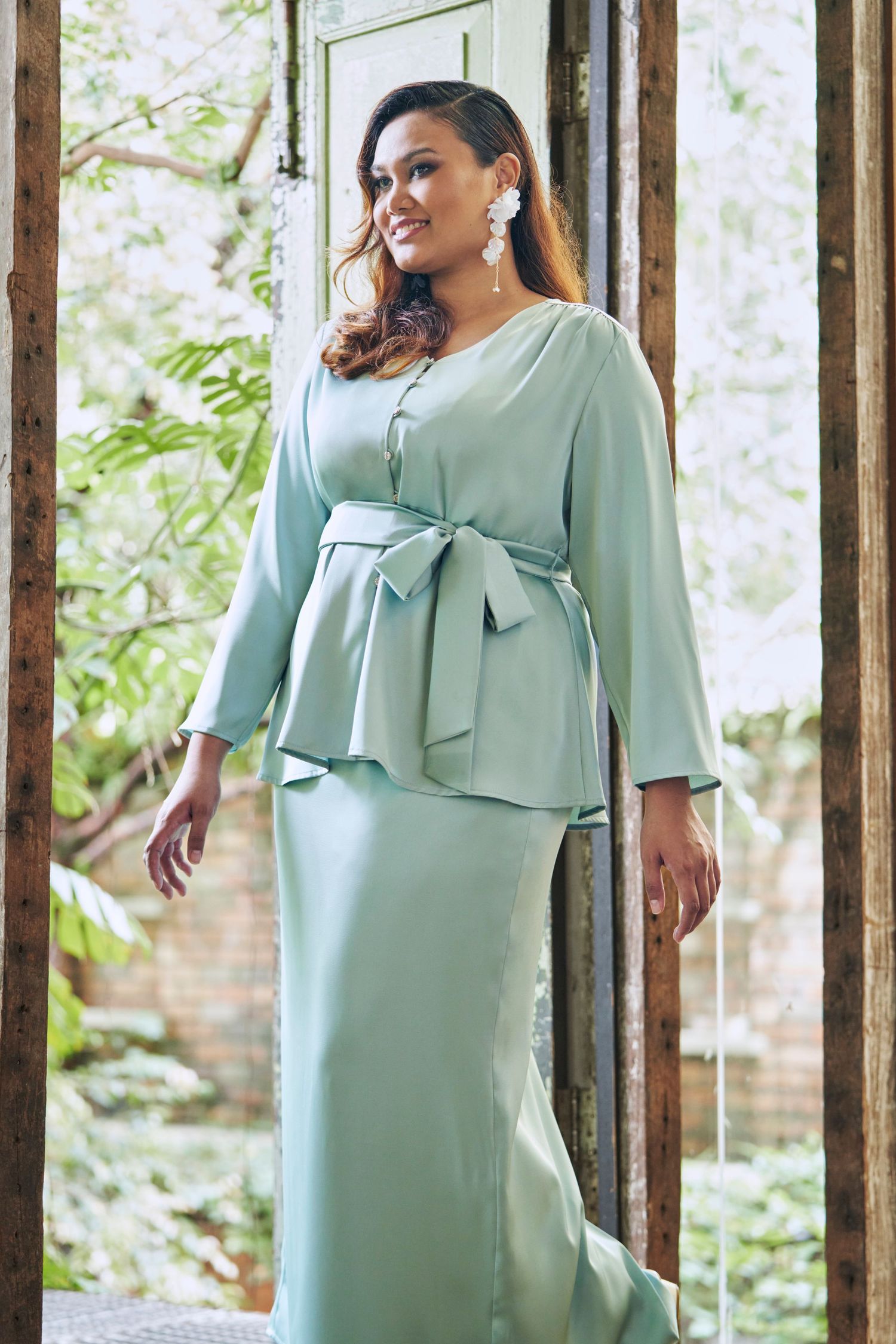Plus-size woman in sage green peplum top and maxi skirt set with bow belt detail.