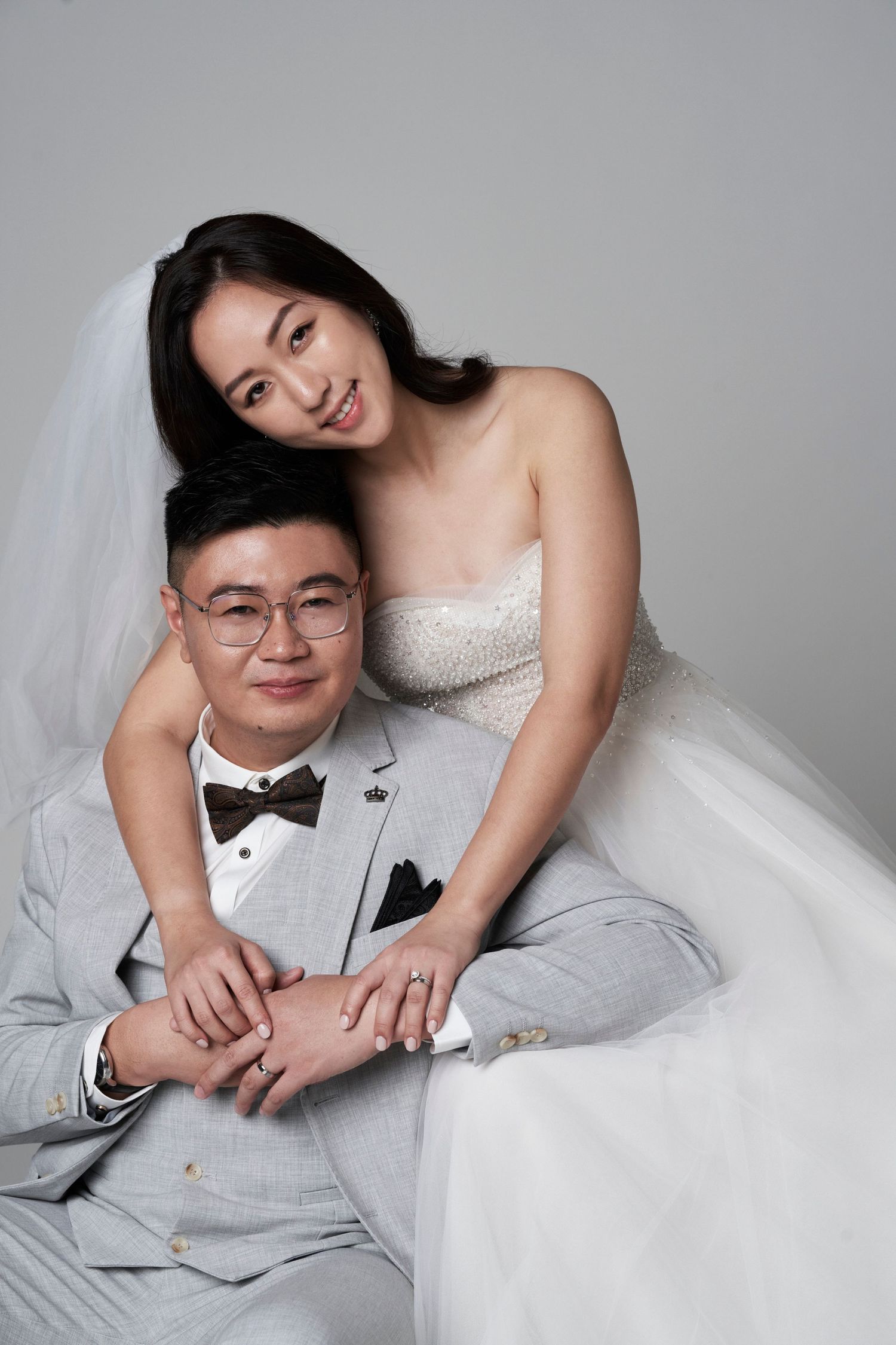 A bride in a strapless white gown leans over her seated groom in glasses during a wedding photo session.