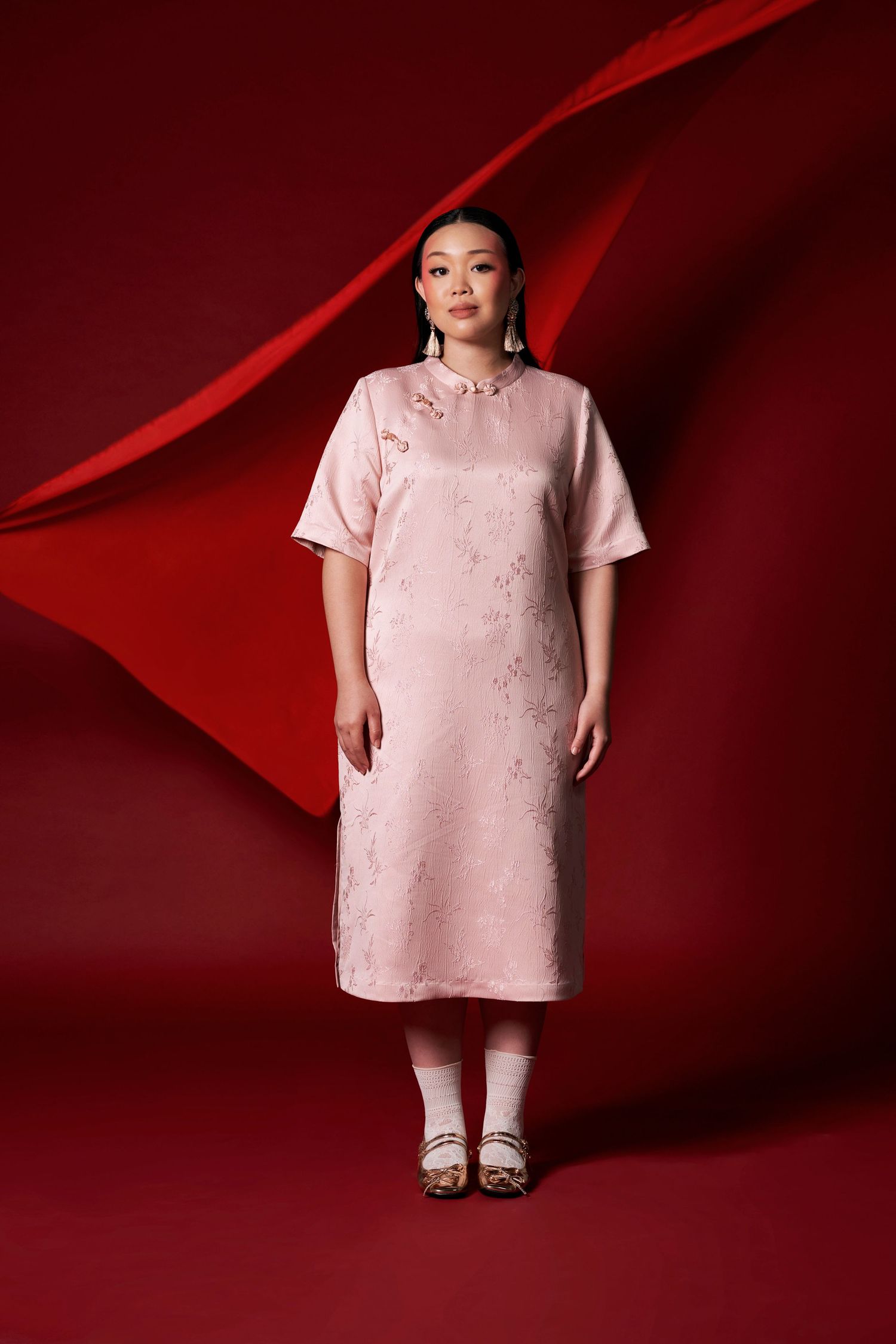 Asian woman in blush pink jacquard cheongsam dress stands confidently against bold red draped background.