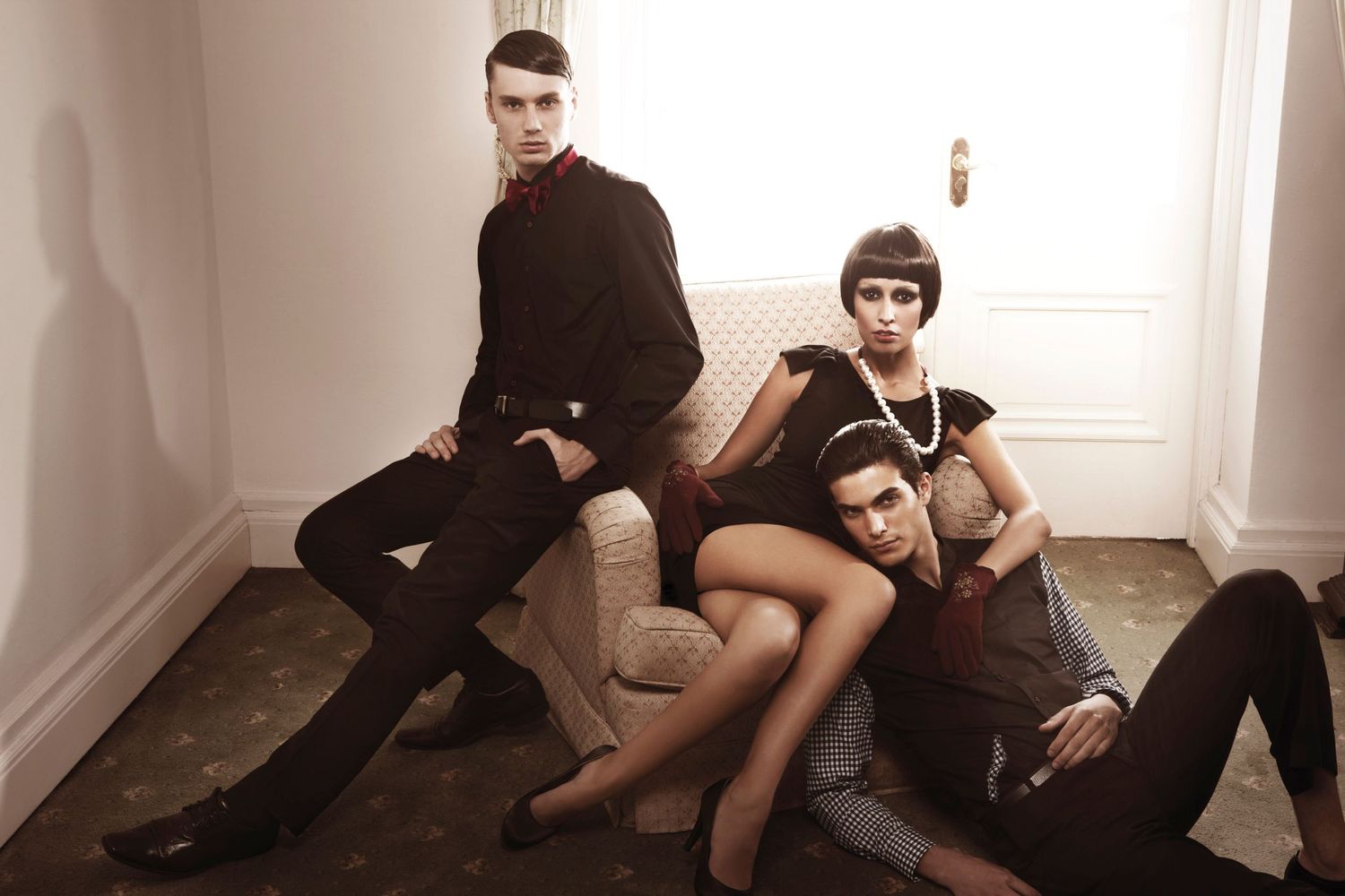 Three fashion models in dark sophisticated outfits pose dramatically on floor and chair in elegant room.