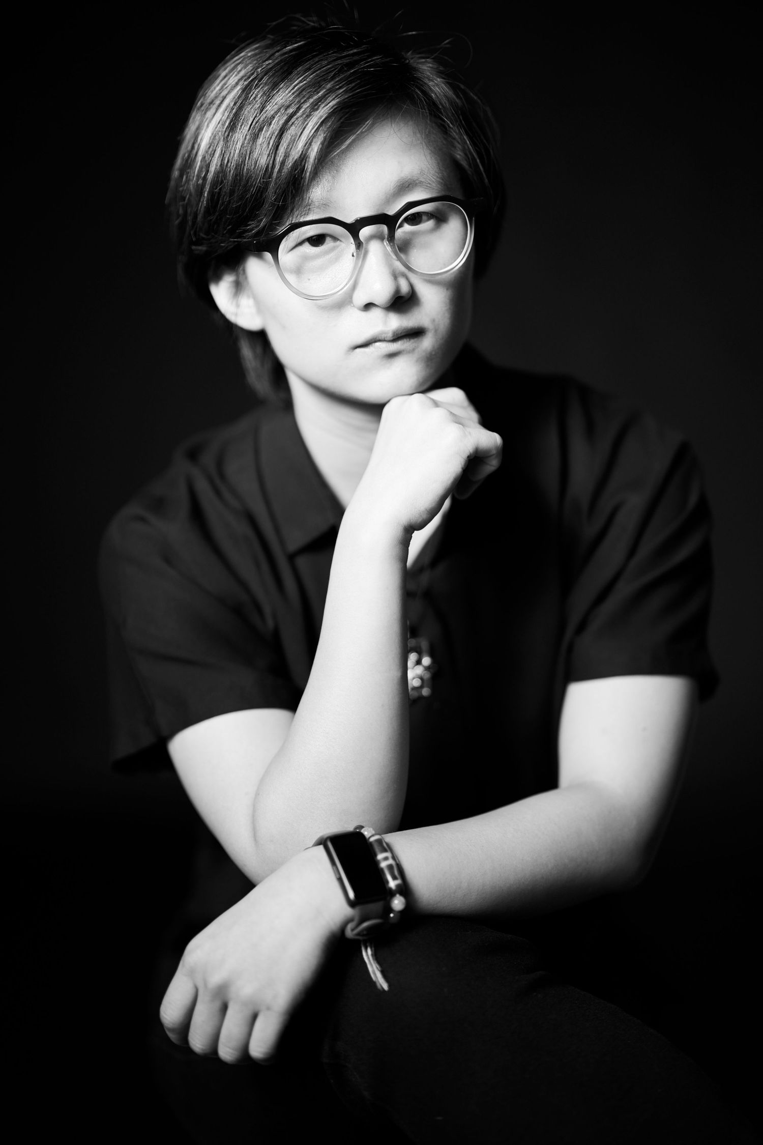 Young person in black polo with glasses poses in black and white studio portrait.