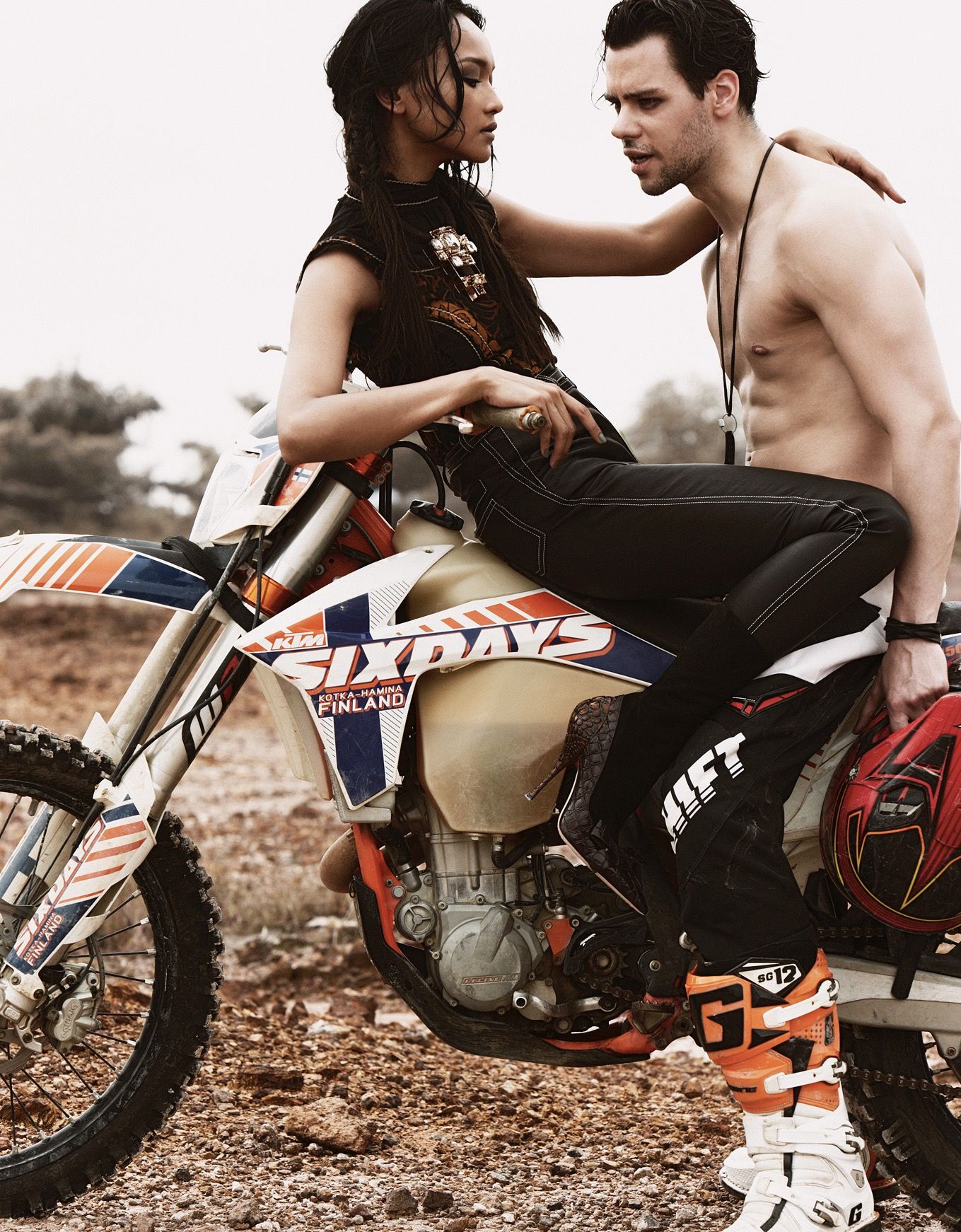 Woman in edgy outfit sits on KTM Six Days dirt bike with shirtless man standing beside her.