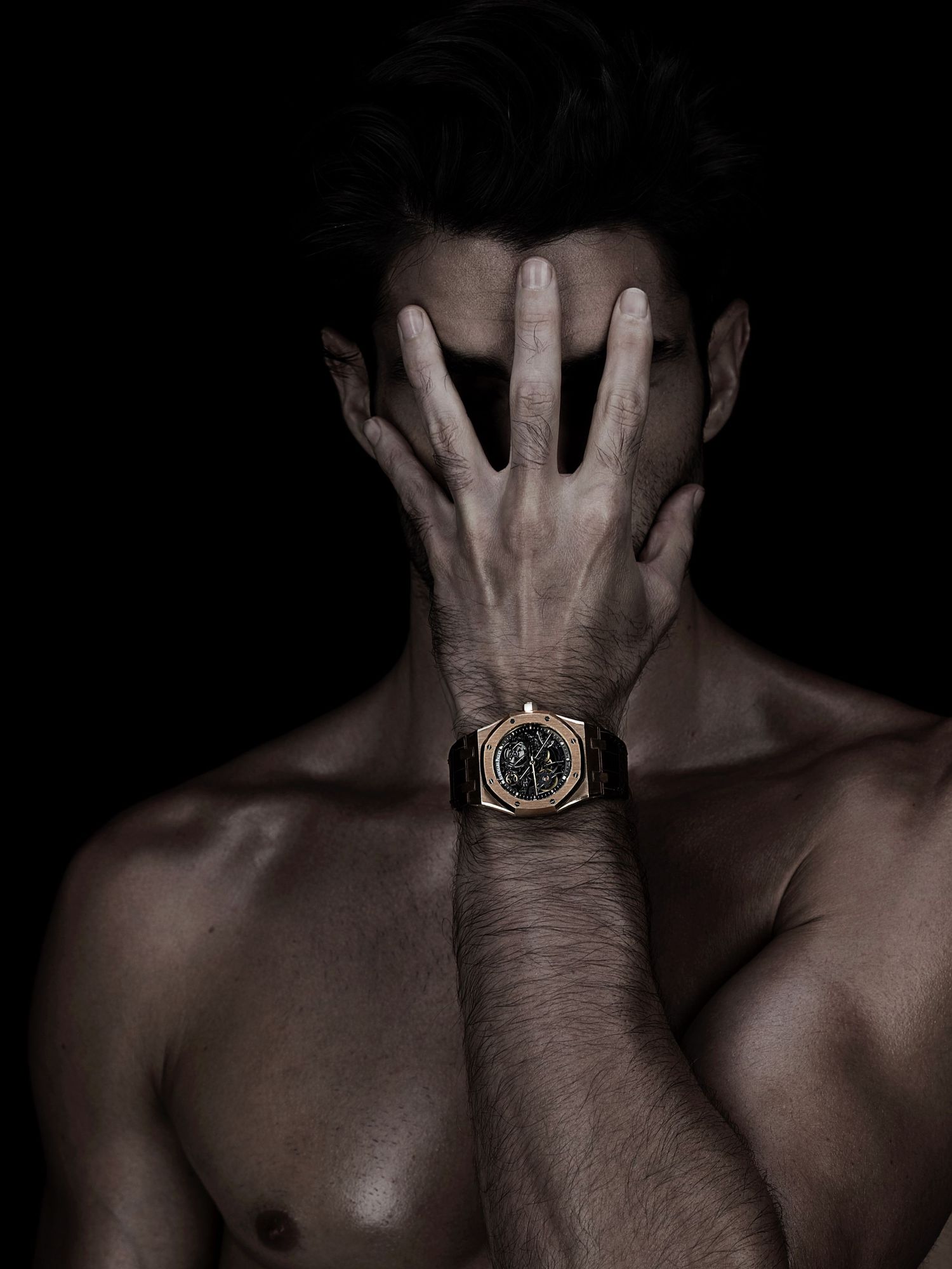 Shirtless man covering face with hands wearing gold luxury watch on dark background.