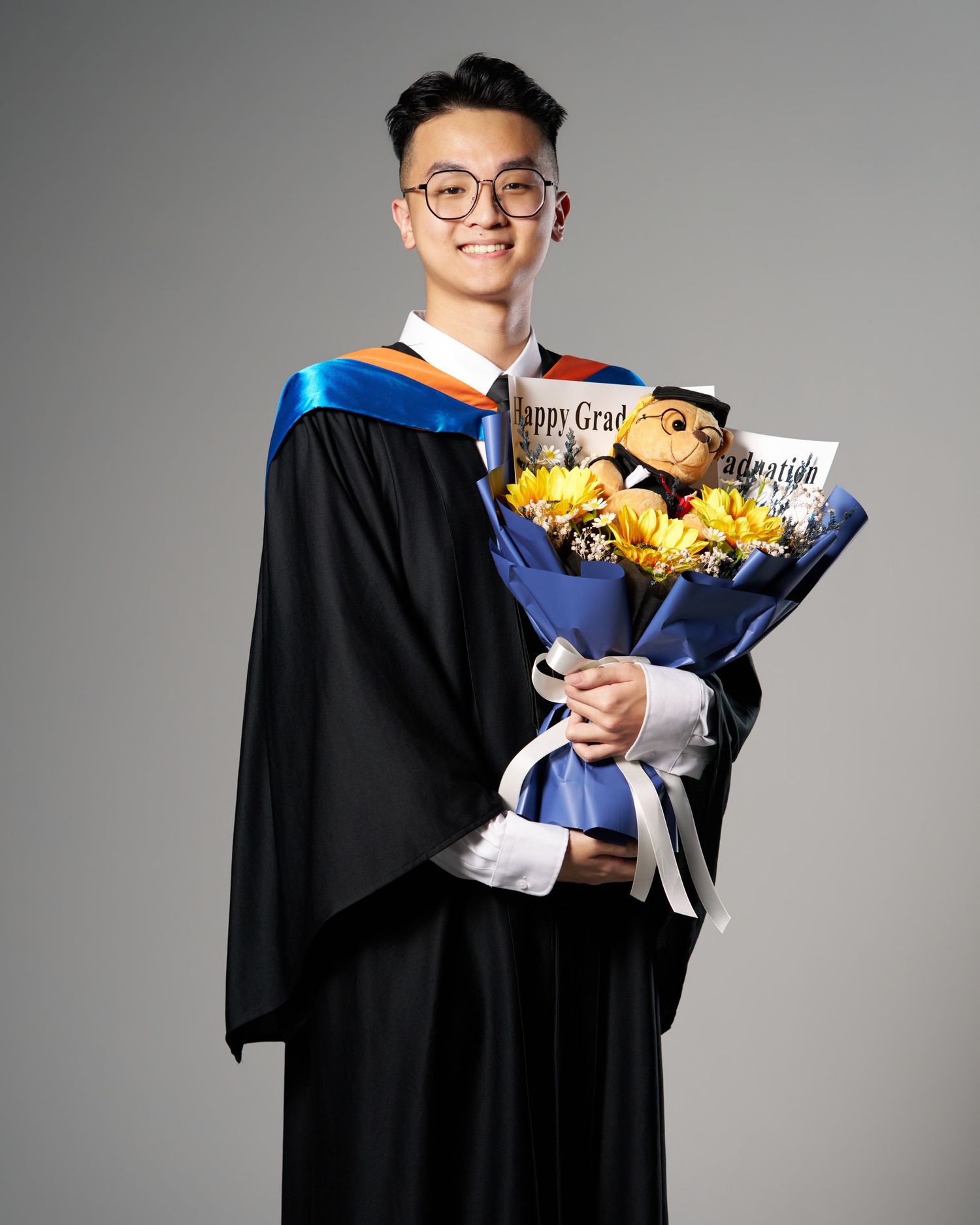 Smiling graduate in black gown with blue sash holding a bouquet of sunflowers.