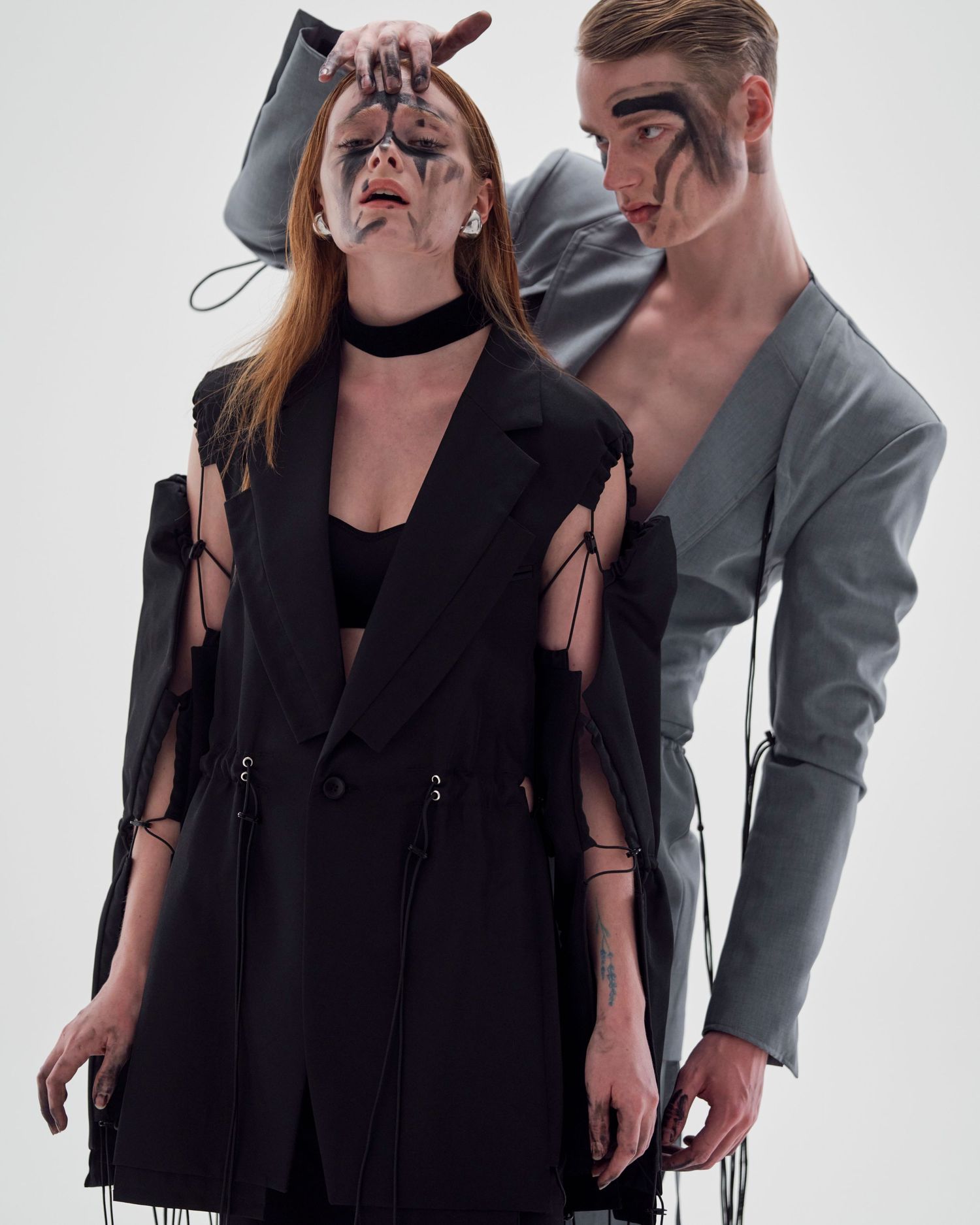 Two models in avant-garde black and gray deconstructed suits with dramatic face paint pose against white background.