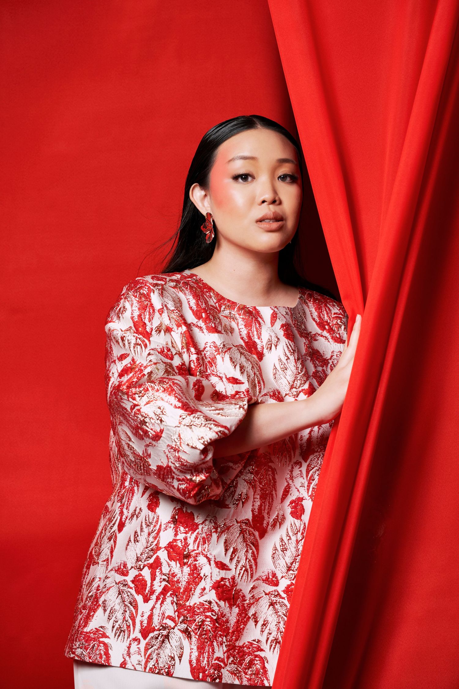 Asian woman in red and white floral print blouse emerges from vibrant red curtain in fashion editorial.