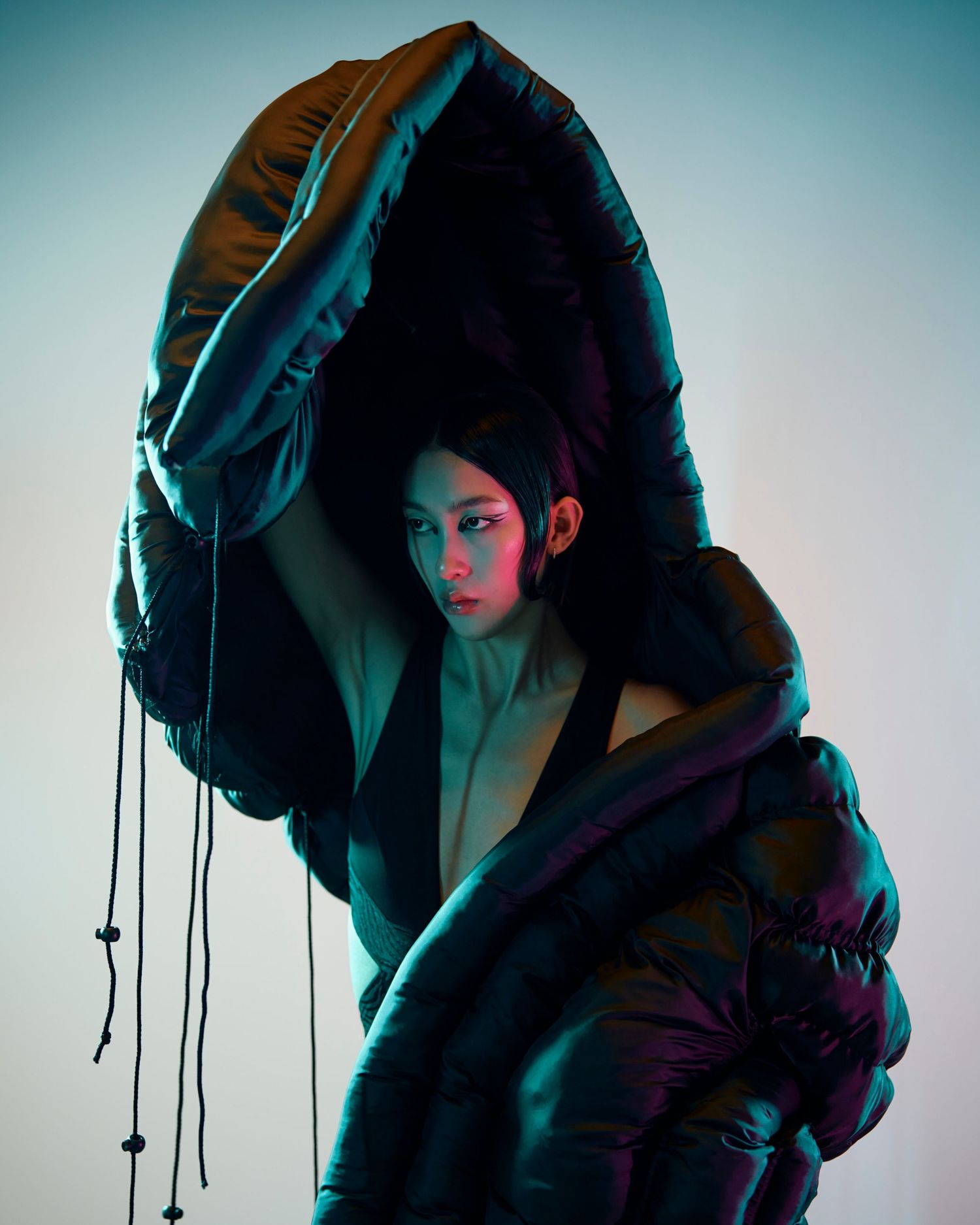 Model draped in large iridescent puffer fabric holding it overhead in artistic fashion editorial shot.