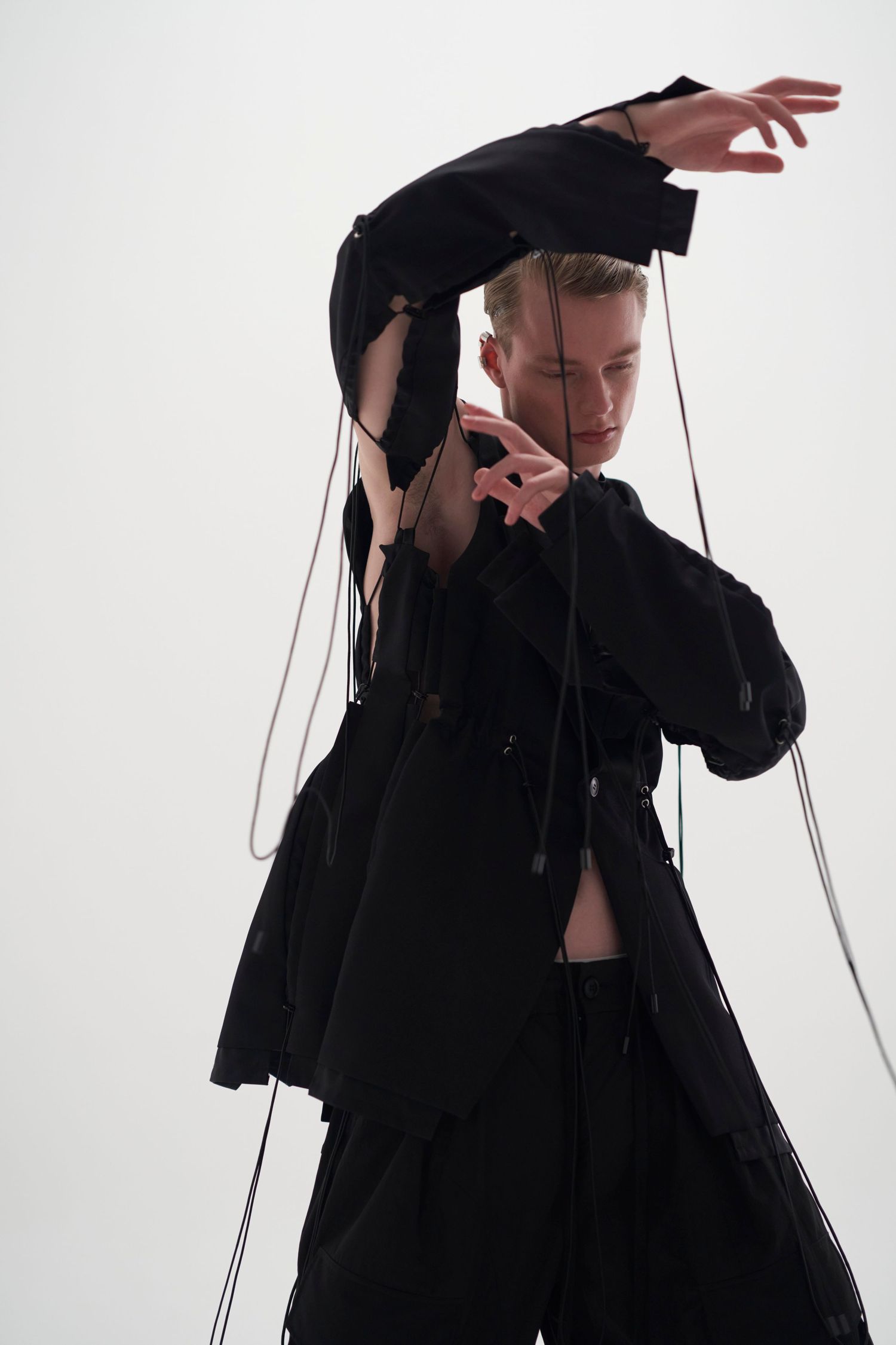 Model in black deconstructed outfit with dangling strings posing dramatically against white background.
