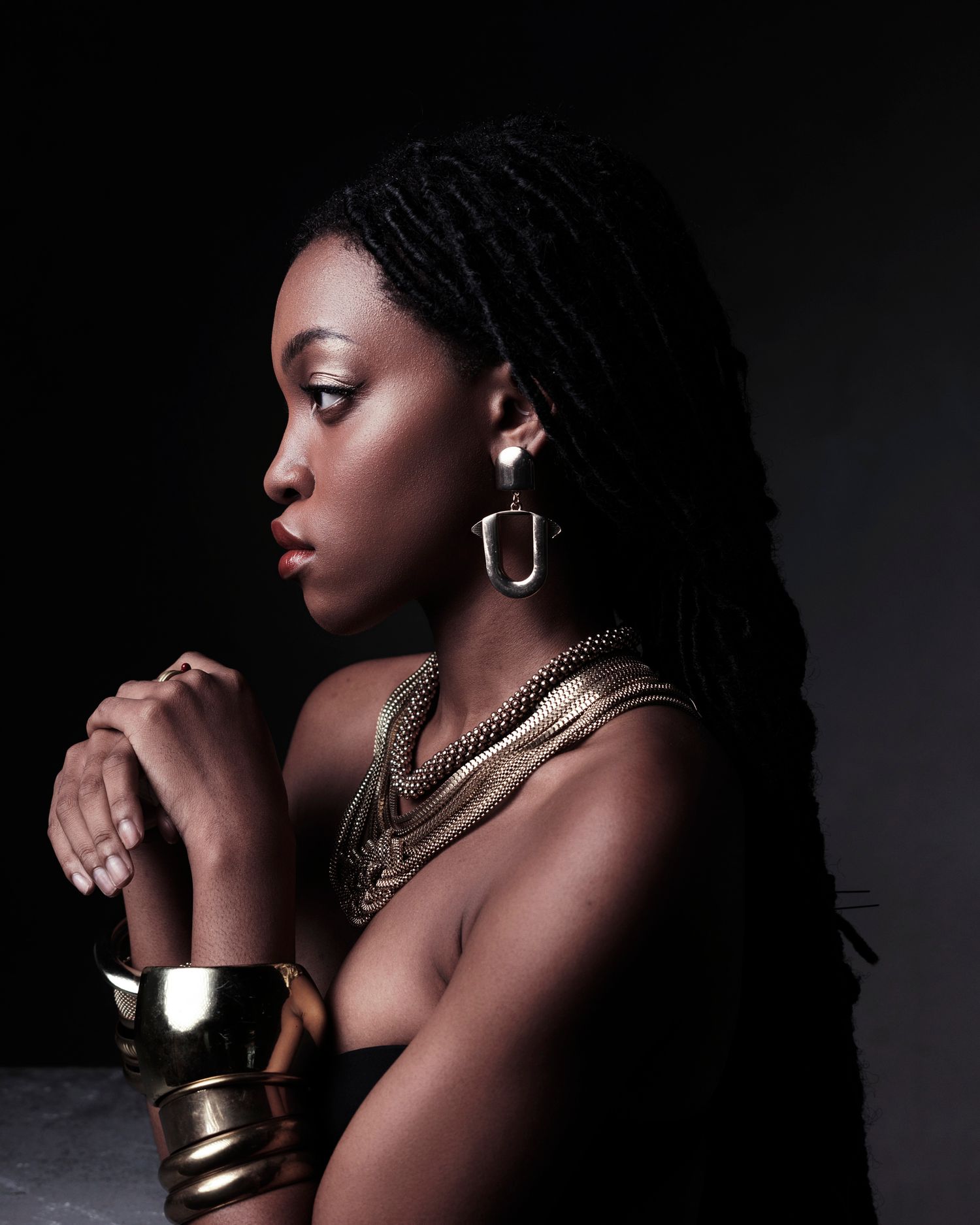 Black woman in profile wearing gold statement jewelry including hoop earrings, layered necklaces, and cuff bracelet.