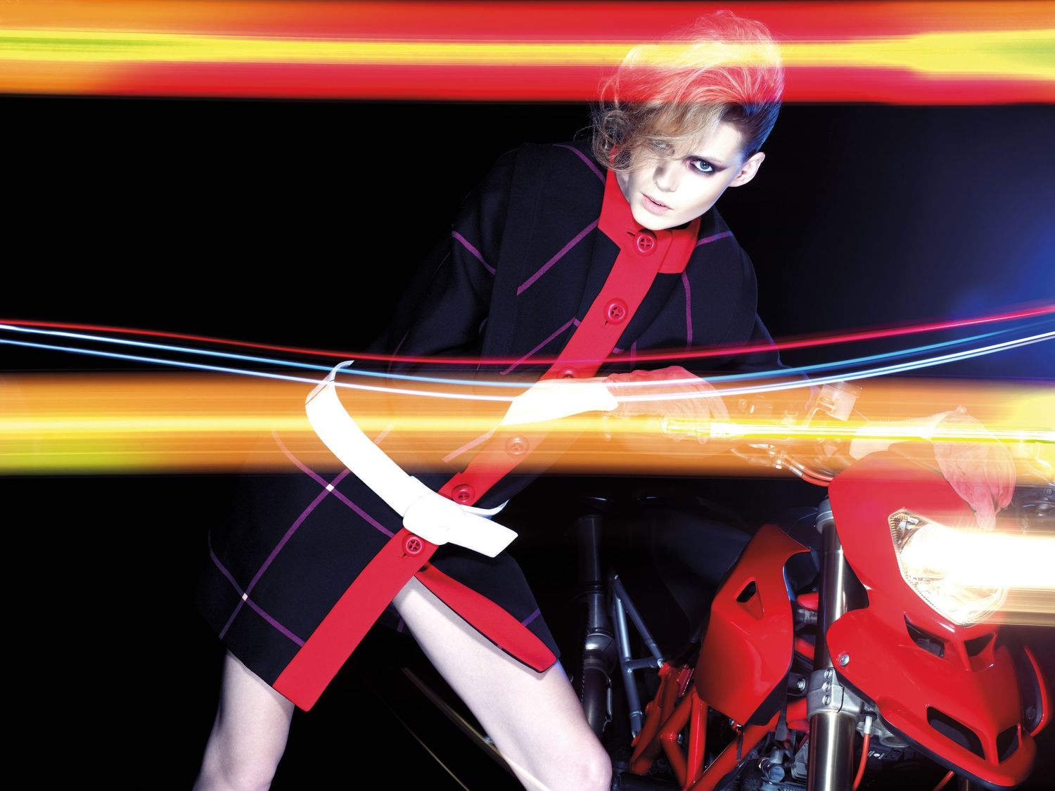 Model in black and red geometric outfit poses dynamically with colorful light trails and motorcycle in fashion editorial.