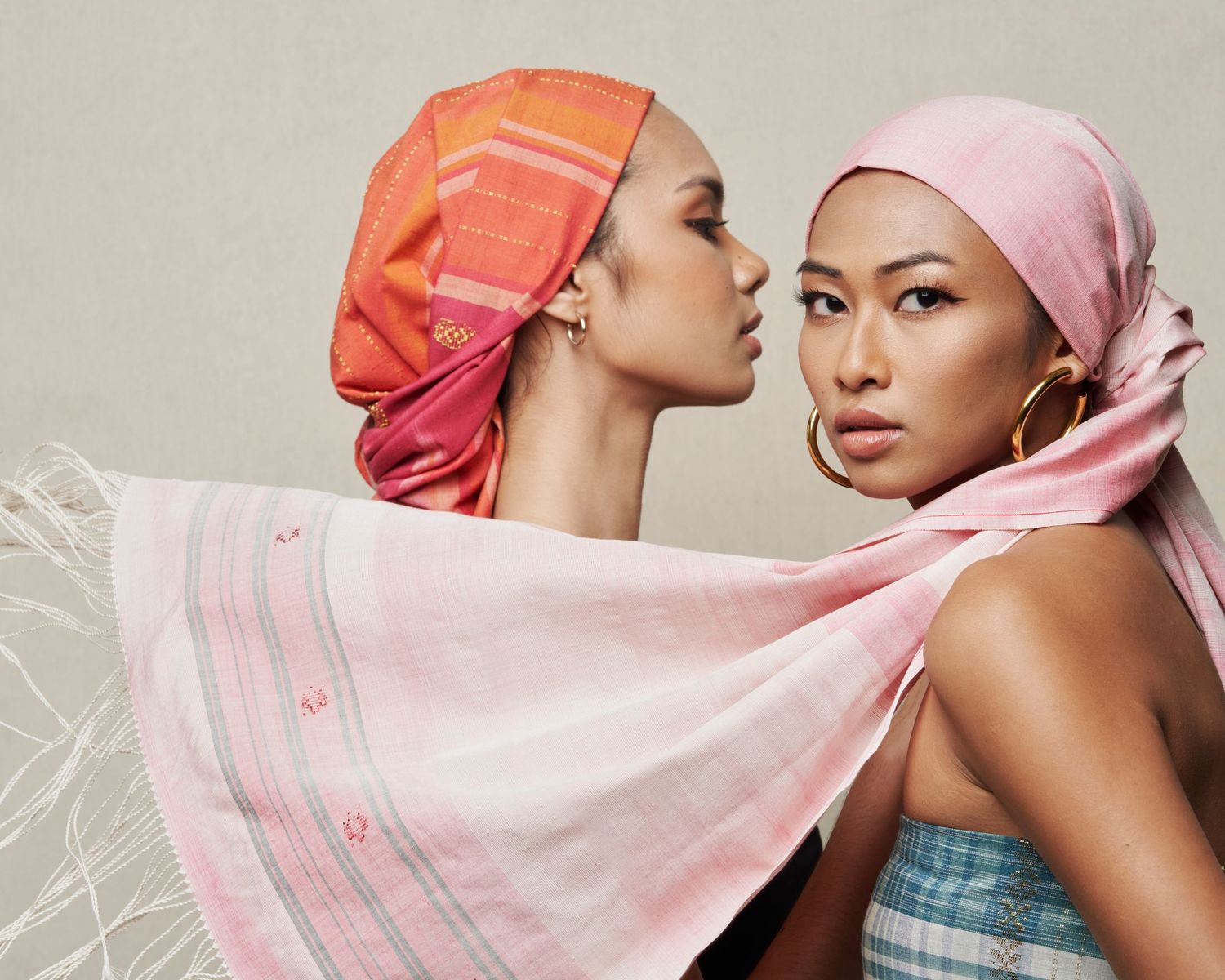 Two women wearing colorful head wraps and pink scarves pose together in a fashion editorial.