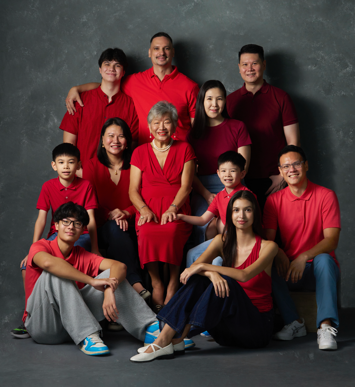 Large multigenerational Asian family portrait with everyone wearing coordinated red outfits on grey background.