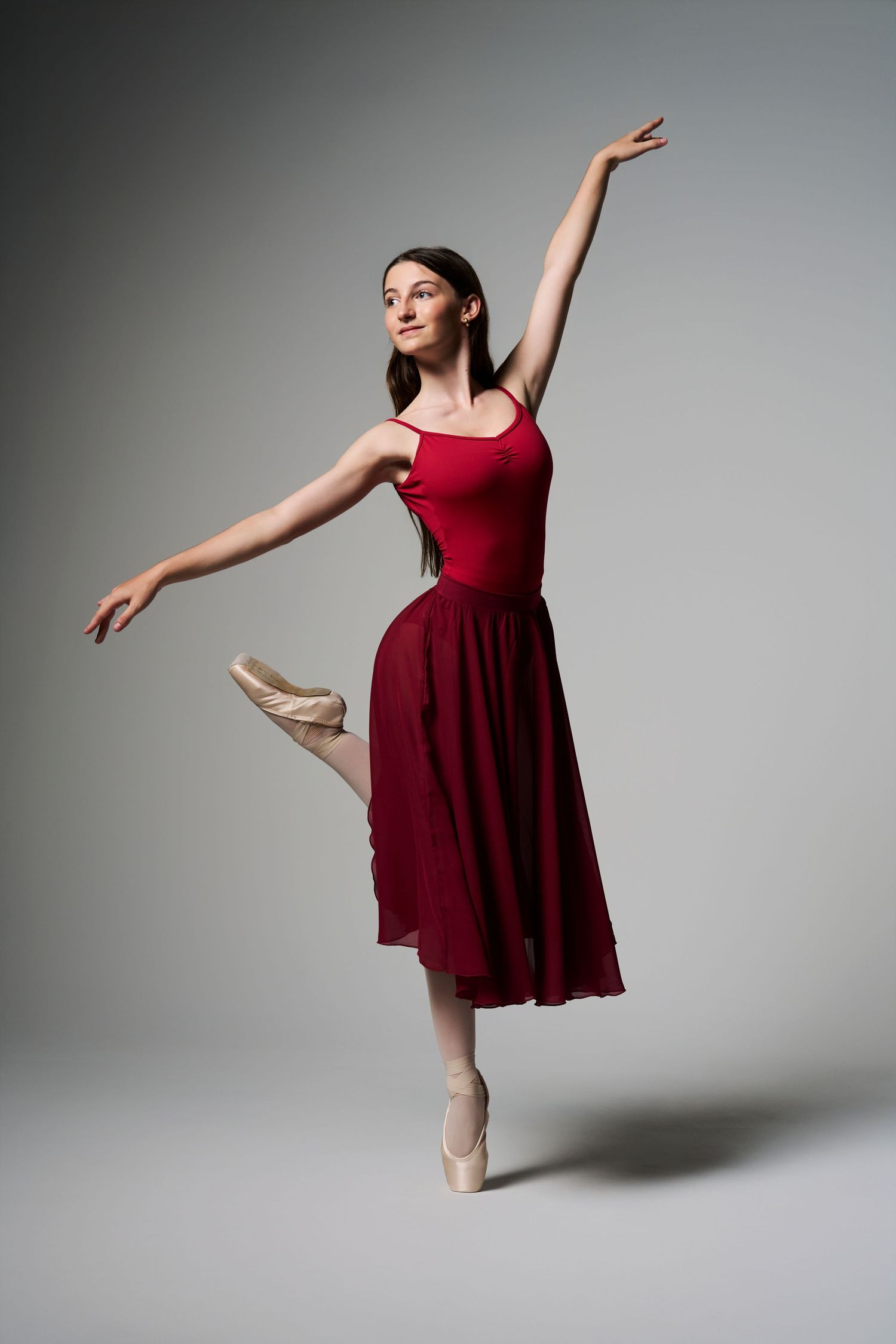 A elegant ballerina poses en pointe in a red dress with arms extended in a gray studio.