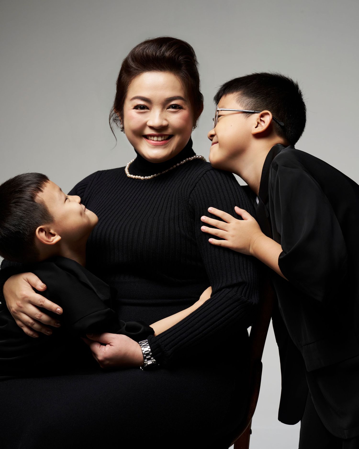 A joyful pregnant woman cuddles with two boys, all dressed in black, in a studio portrait.