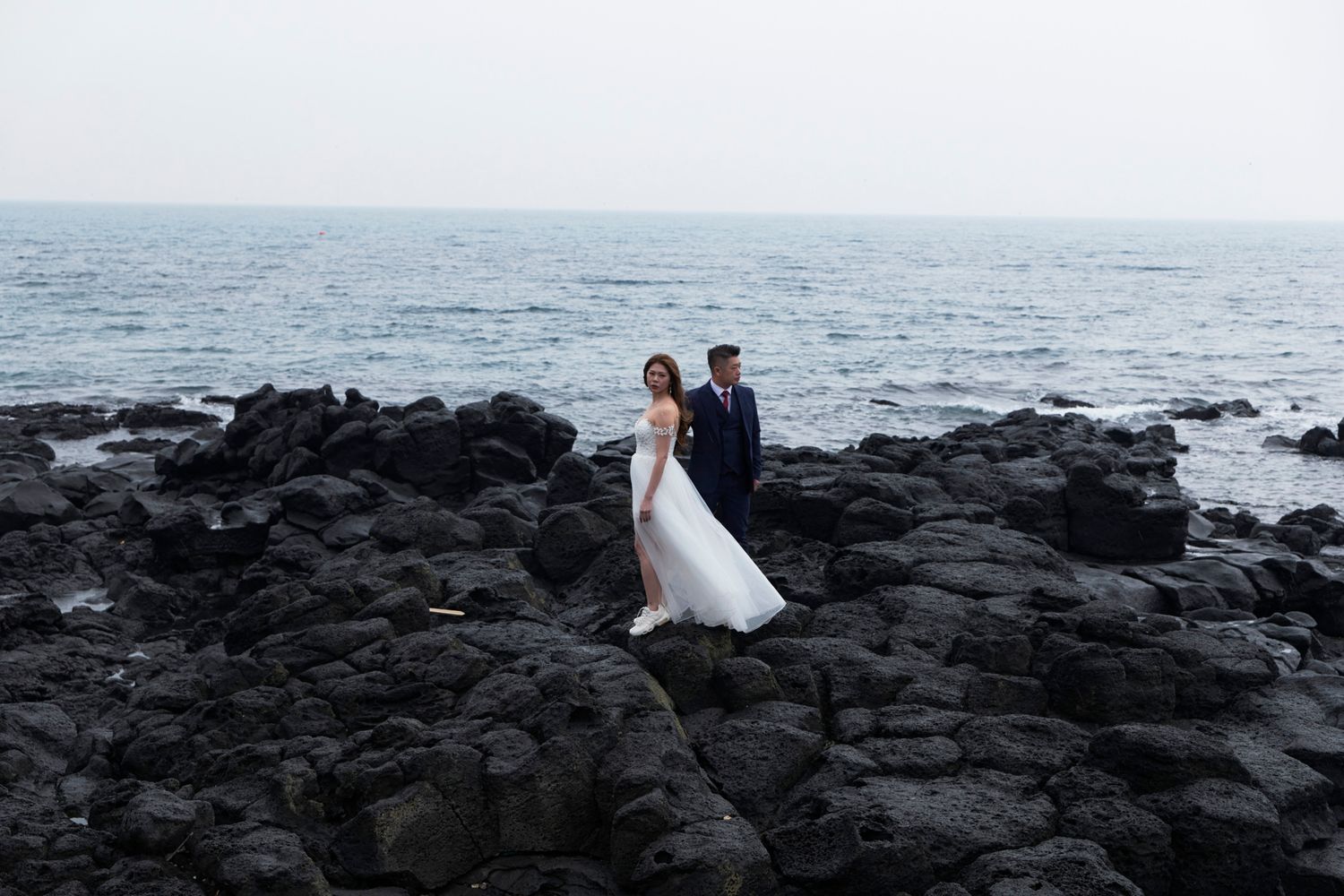 Bride and groom standing together on rugged black volcanic rocks beside the ocean.