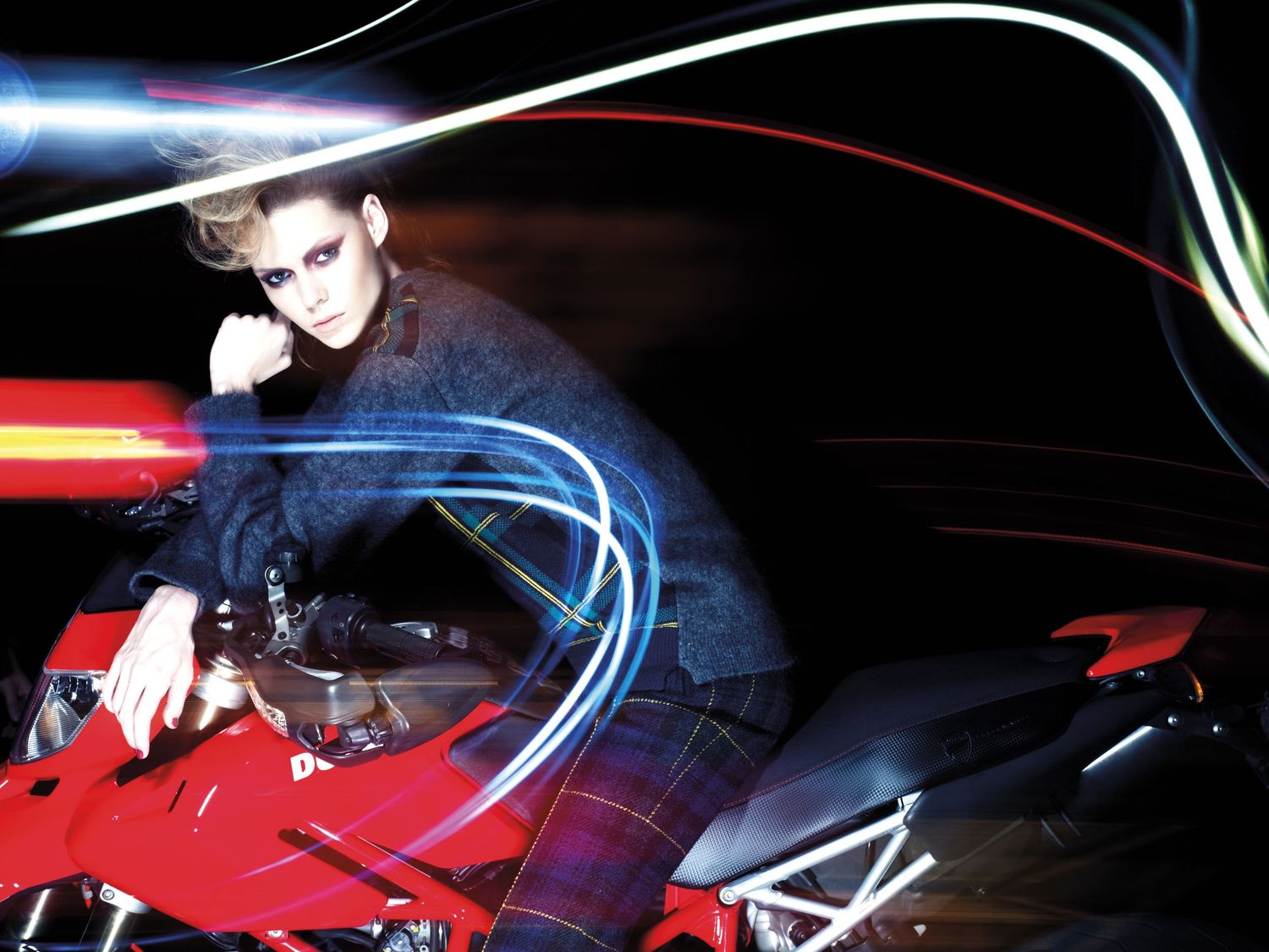 Female model in dark outfit poses on red Ducati motorcycle with colorful light trails.