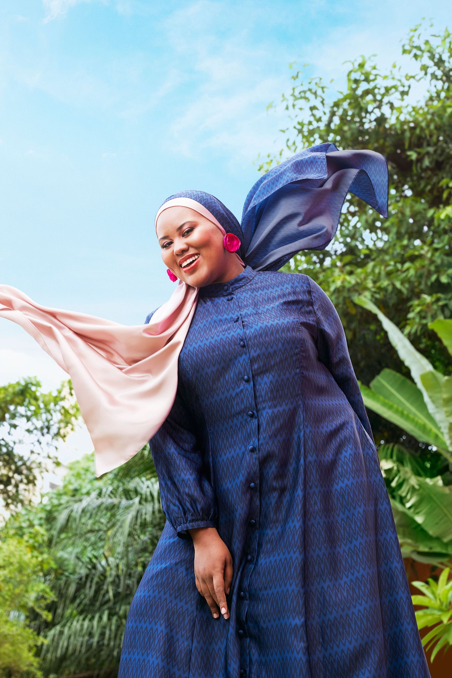 Plus-size Muslim woman in navy blue button-down dress with flowing pink hijab outdoors.