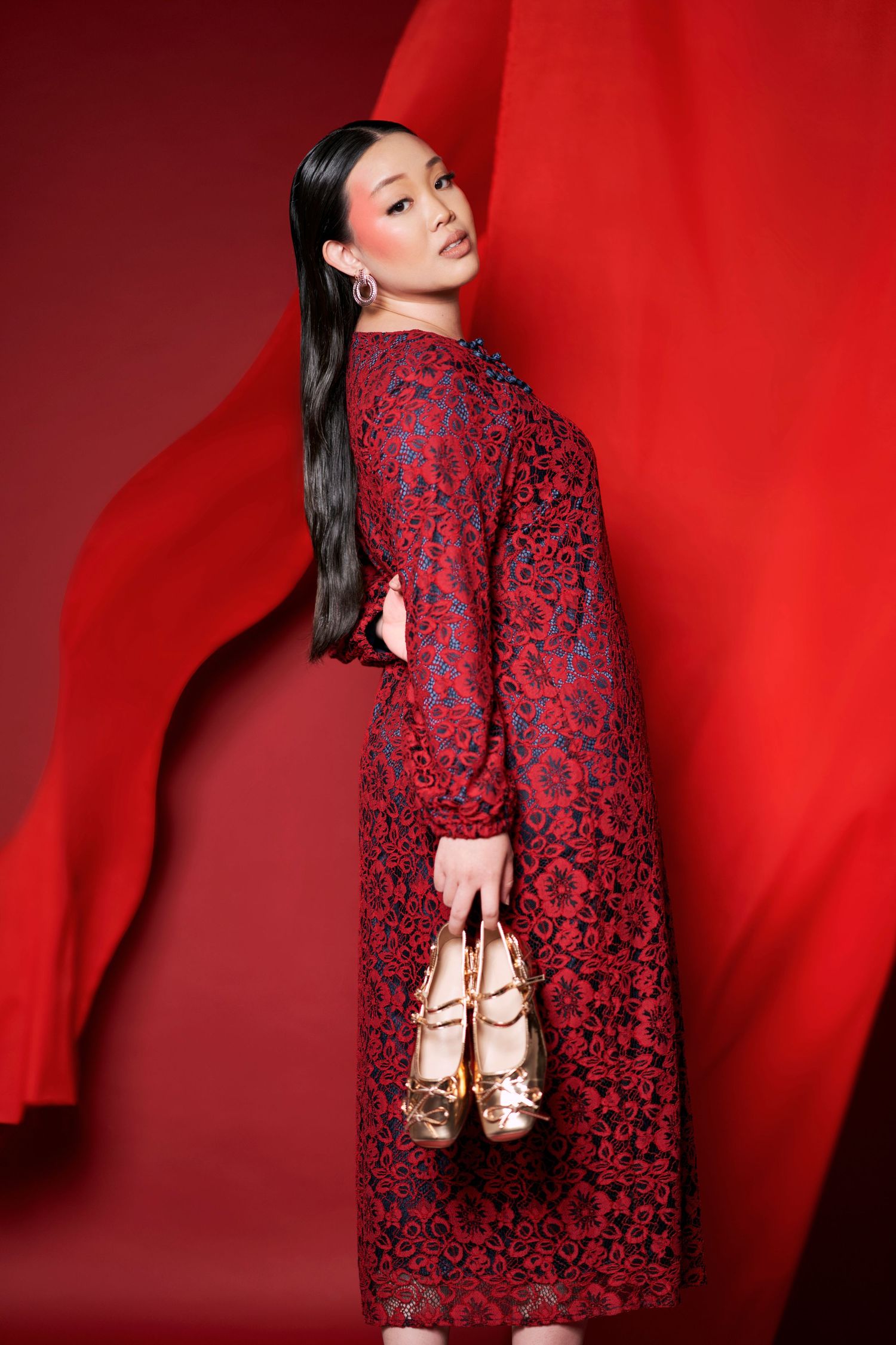 Asian woman in red floral lace midi dress holds gold heels against vibrant red draped background.