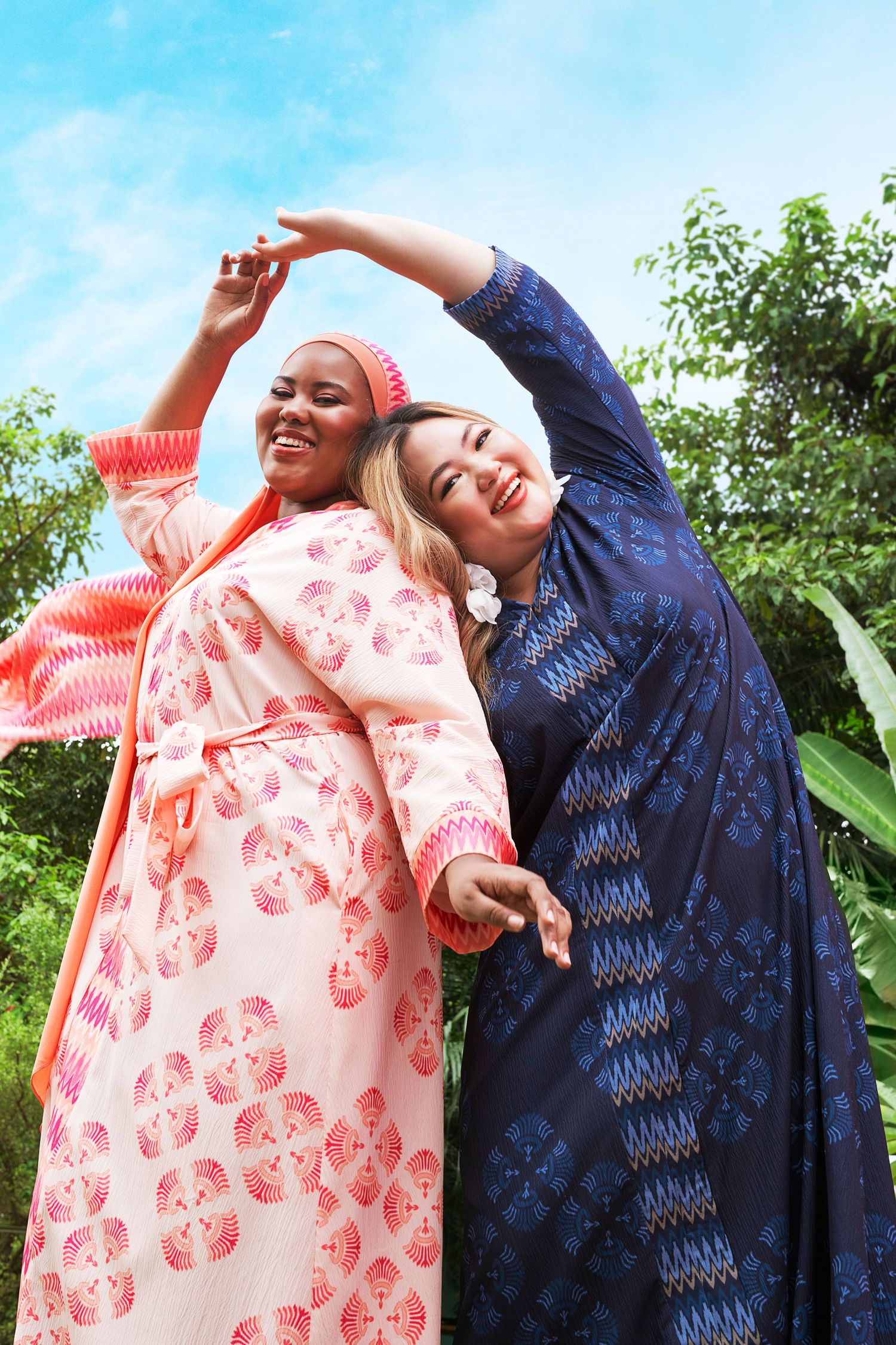 Two plus-size women in colorful printed maxi dresses dancing together in a tropical garden.