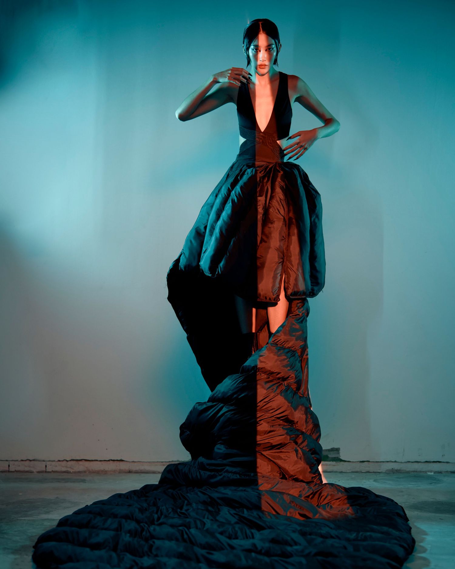 Model in dramatic deep-plunge black and brown sculptural gown with voluminous train under teal lighting.