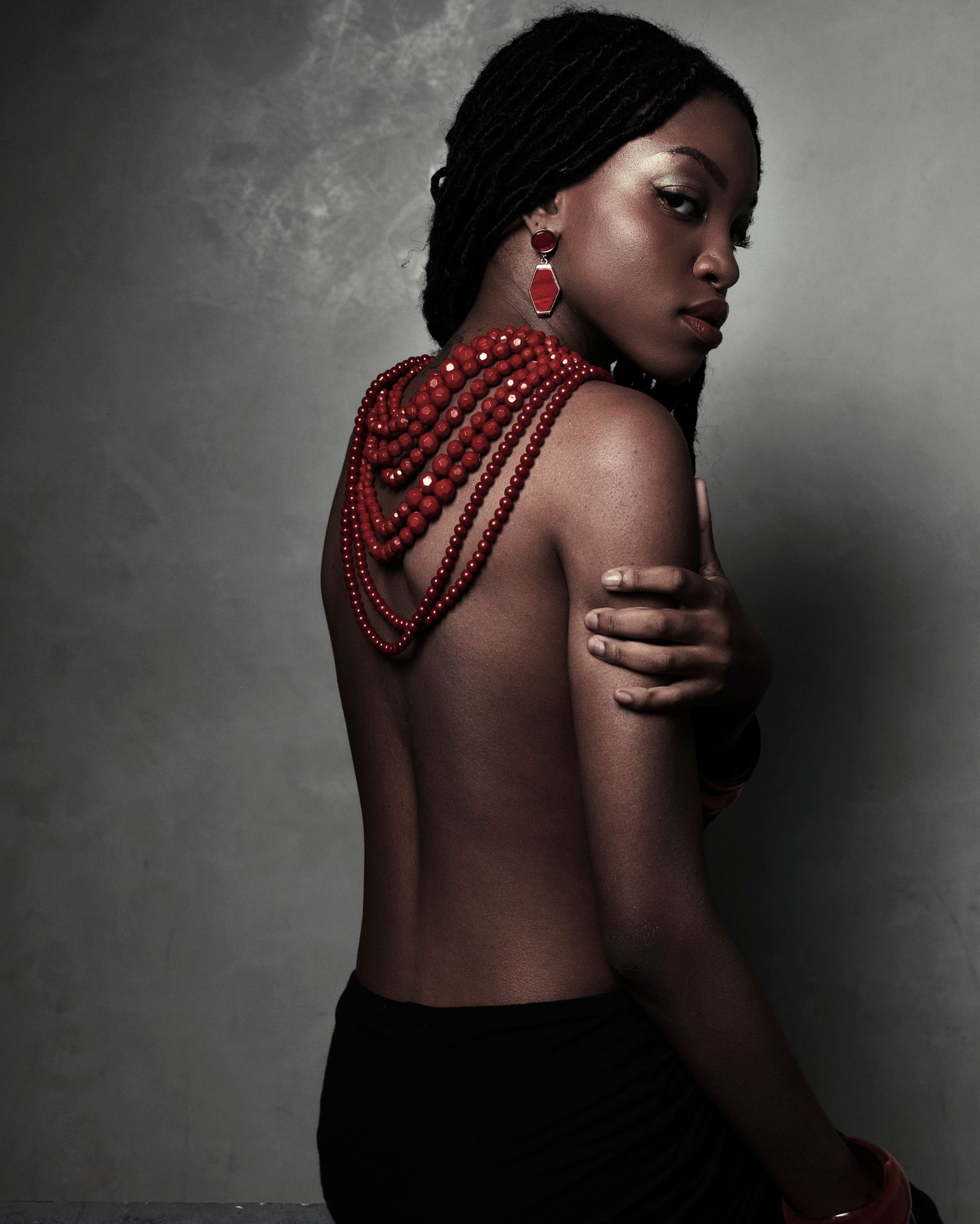 Black woman with red beaded shoulder necklace and earrings posing with bare back against gray background.