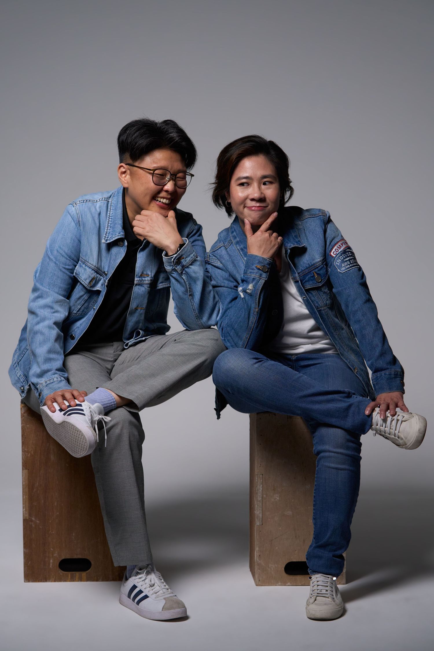 Two smiling Asian people in denim jackets sit on wooden boxes with hands on chins in a studio.
