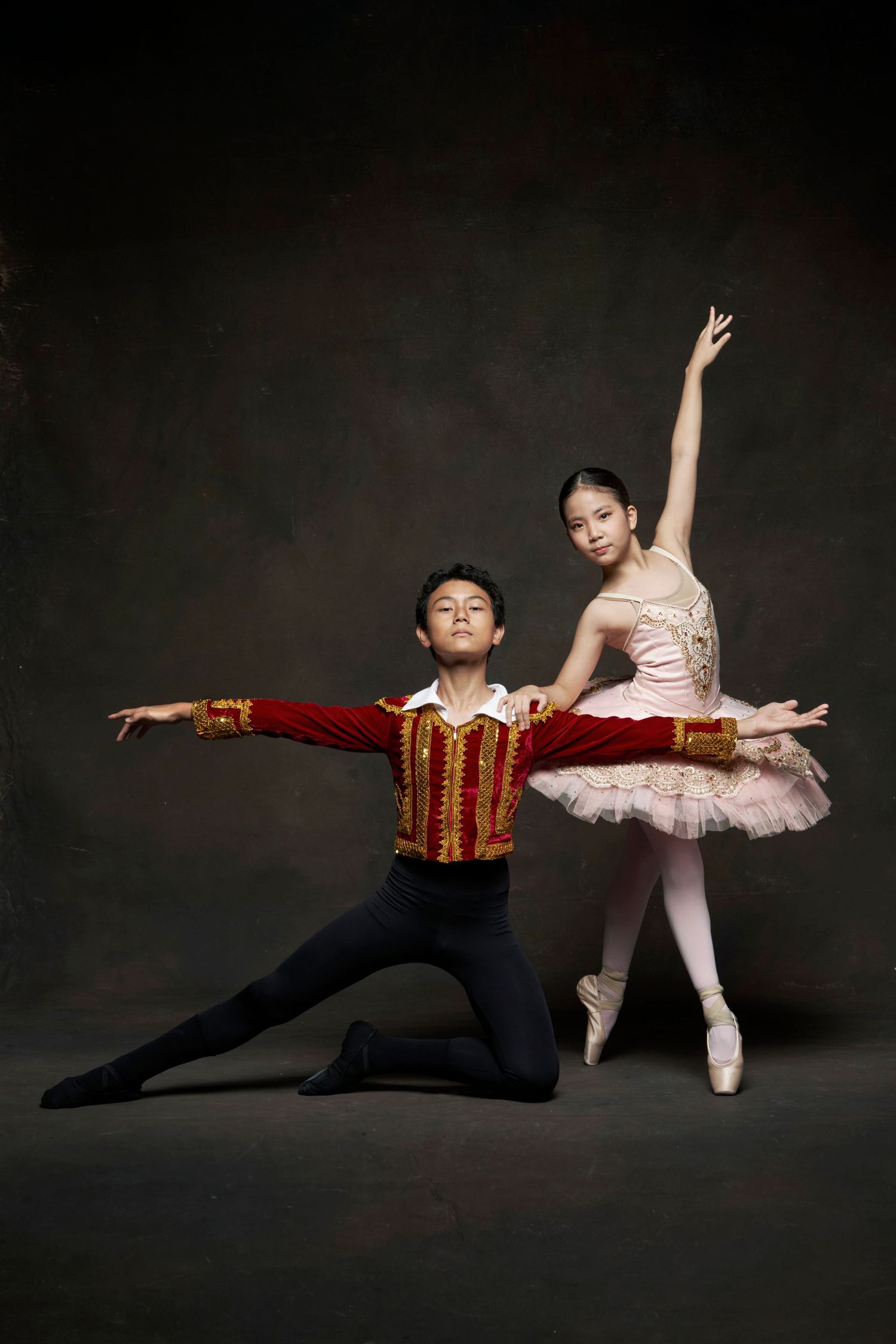 Two young ballet dancers in costume performing classical pas de deux against dark studio backdrop.